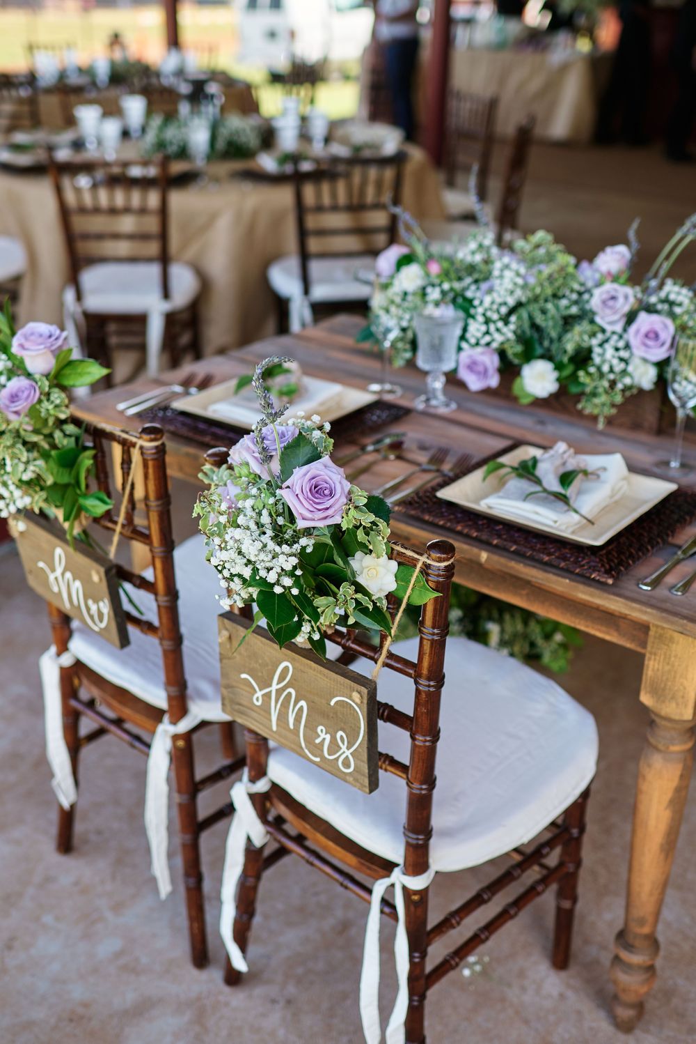 Reception table settings with purple floral arrangements and wooden chairs.