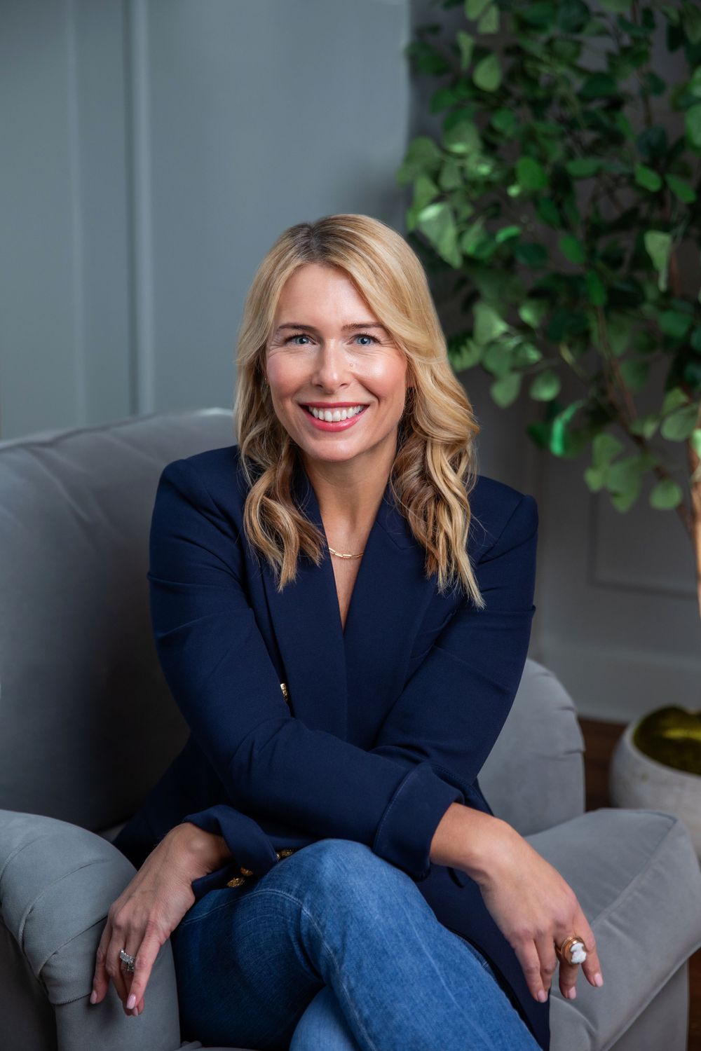 Business professional in navy jacket poses confidently on gray couch with greenery behind.