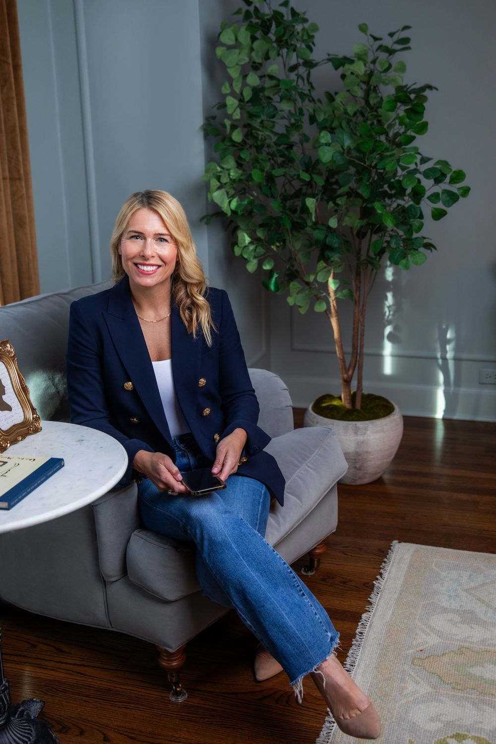 Professional woman in navy blazer sits casually in gray armchair with potted plant in background.