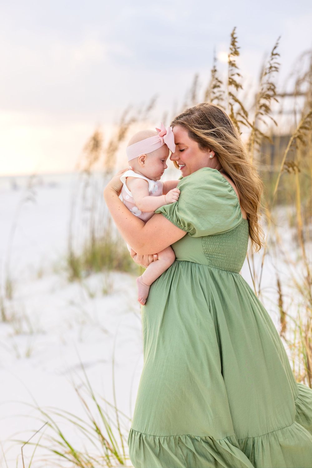 A mother in a sage green dress holds a baby against a beach backdrop with sea oats at sunset.