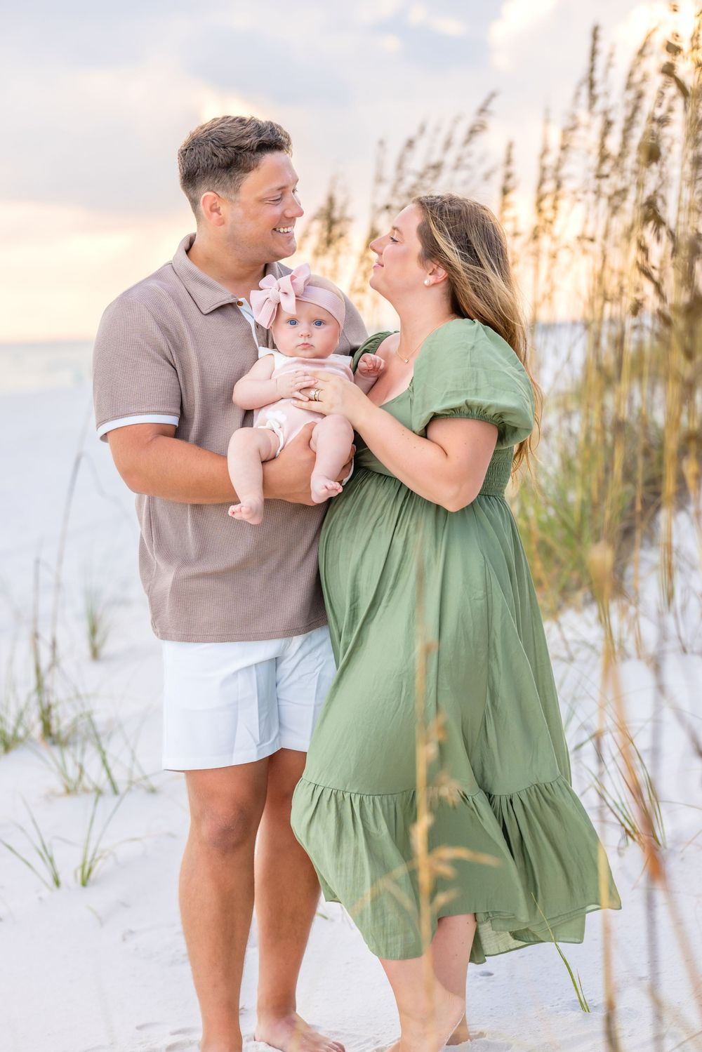A family in coordinated outfits shares tender moments together on a white sandy beach at sunset with sea oats in background.