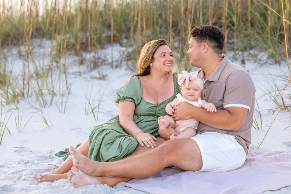 A couple sits together with their baby on a white sandy beach surrounded by sea grass at sunset.