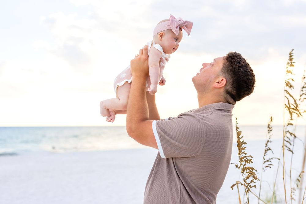 A couple in white attire shares a romantic moment on a sunny beach with sea oats swaying in the breeze.