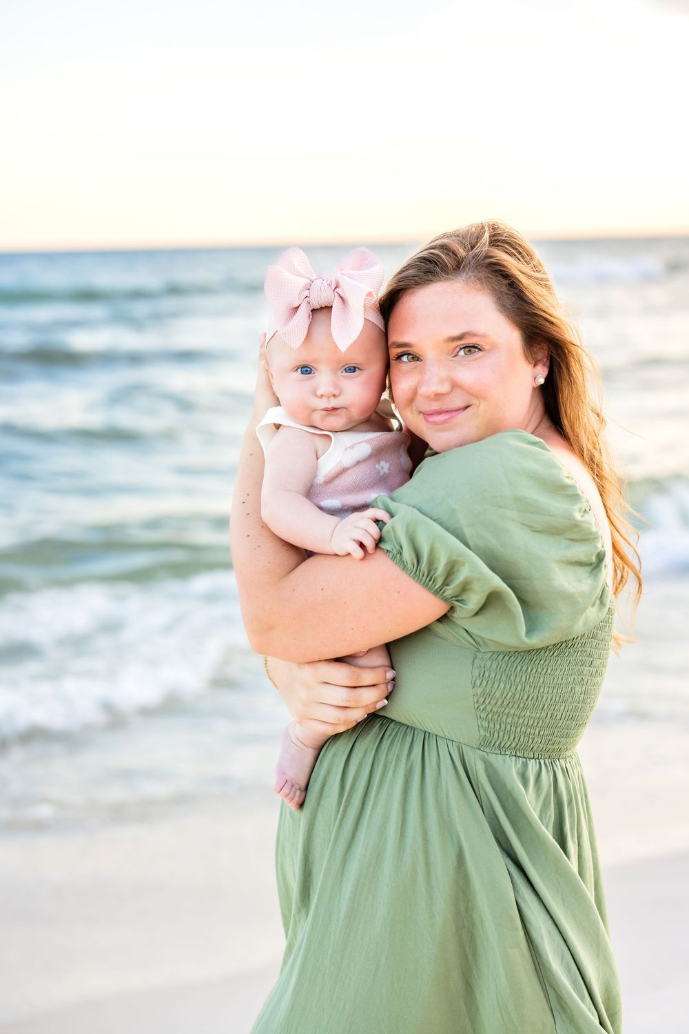 A parent in a sage green dress holds a baby against the ocean waves at sunset.