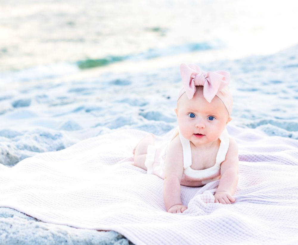 Baby in white outfit with pink bow lies on blanket at beach with soft sand and ocean in background.