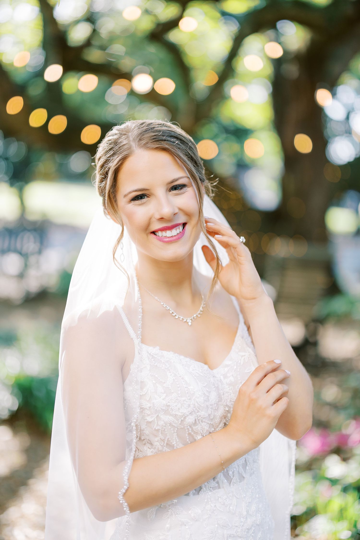 A bride in a white lace dress smiles radiantly in a garden setting with twinkling lights in the background.