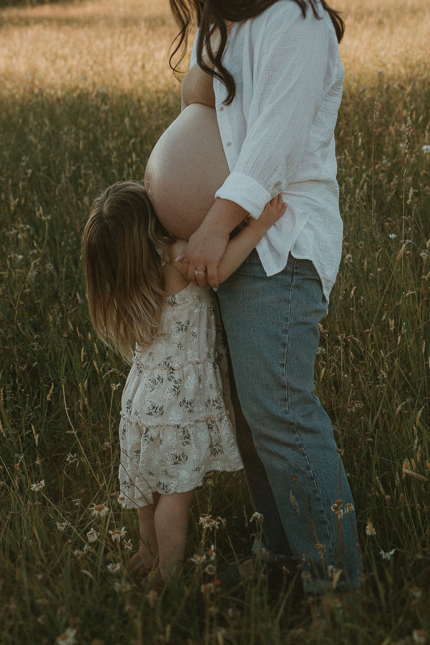 A romantic couple embraces in a sunlit meadow with tall grass, wearing casual white and denim outfits.