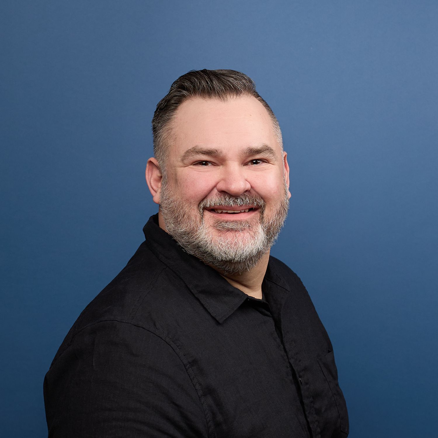 Headshot portrait of man with black hair and black shirt smiling with blue background