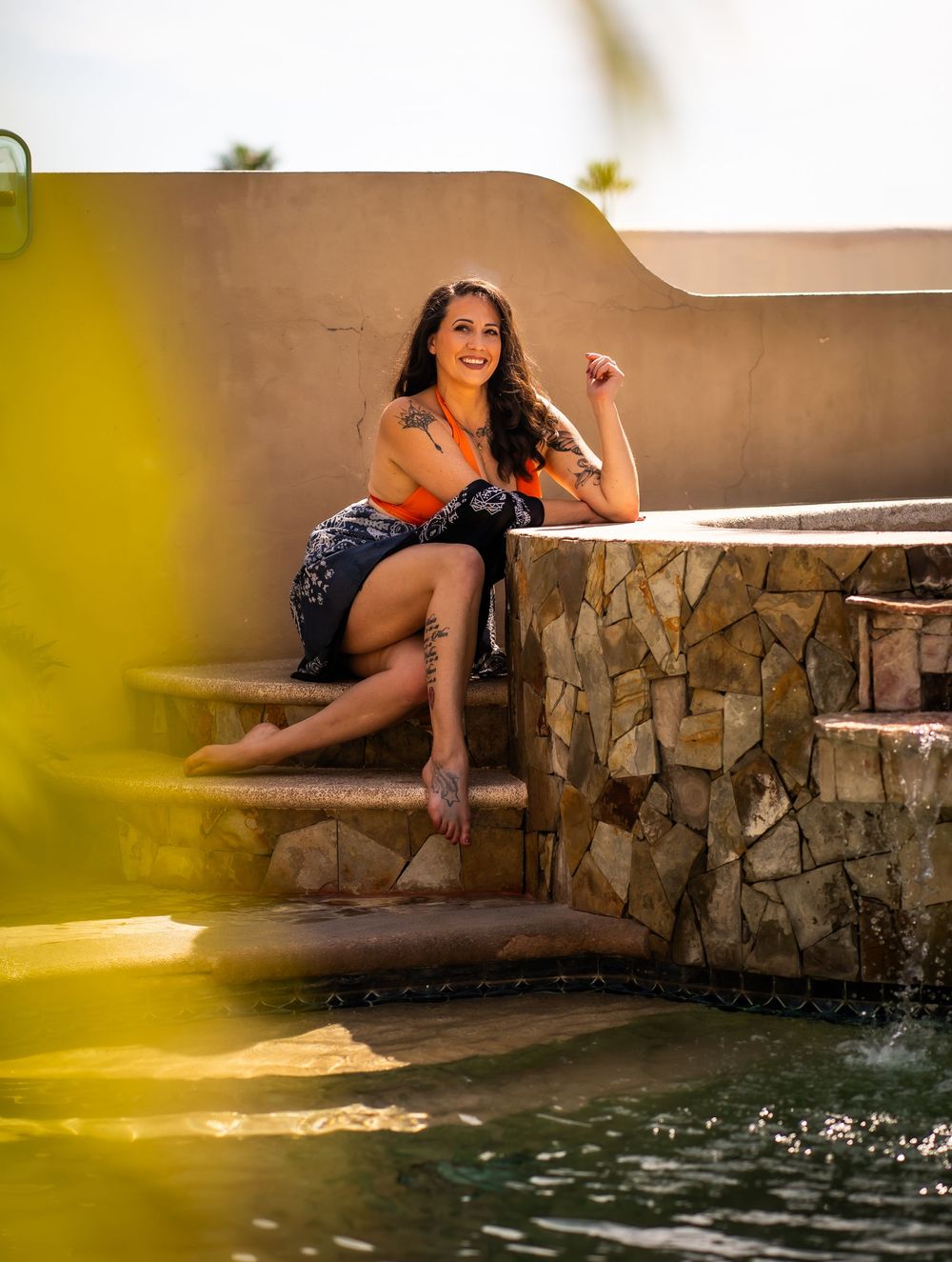 Woman in casual attire sits on stone steps by a pool during golden hour sunlight.