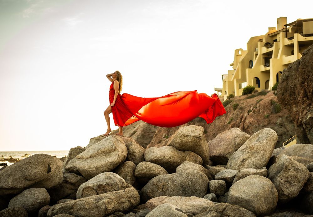 A flowing red fabric creates an artistic movement across a rocky coastal landscape at sunset.