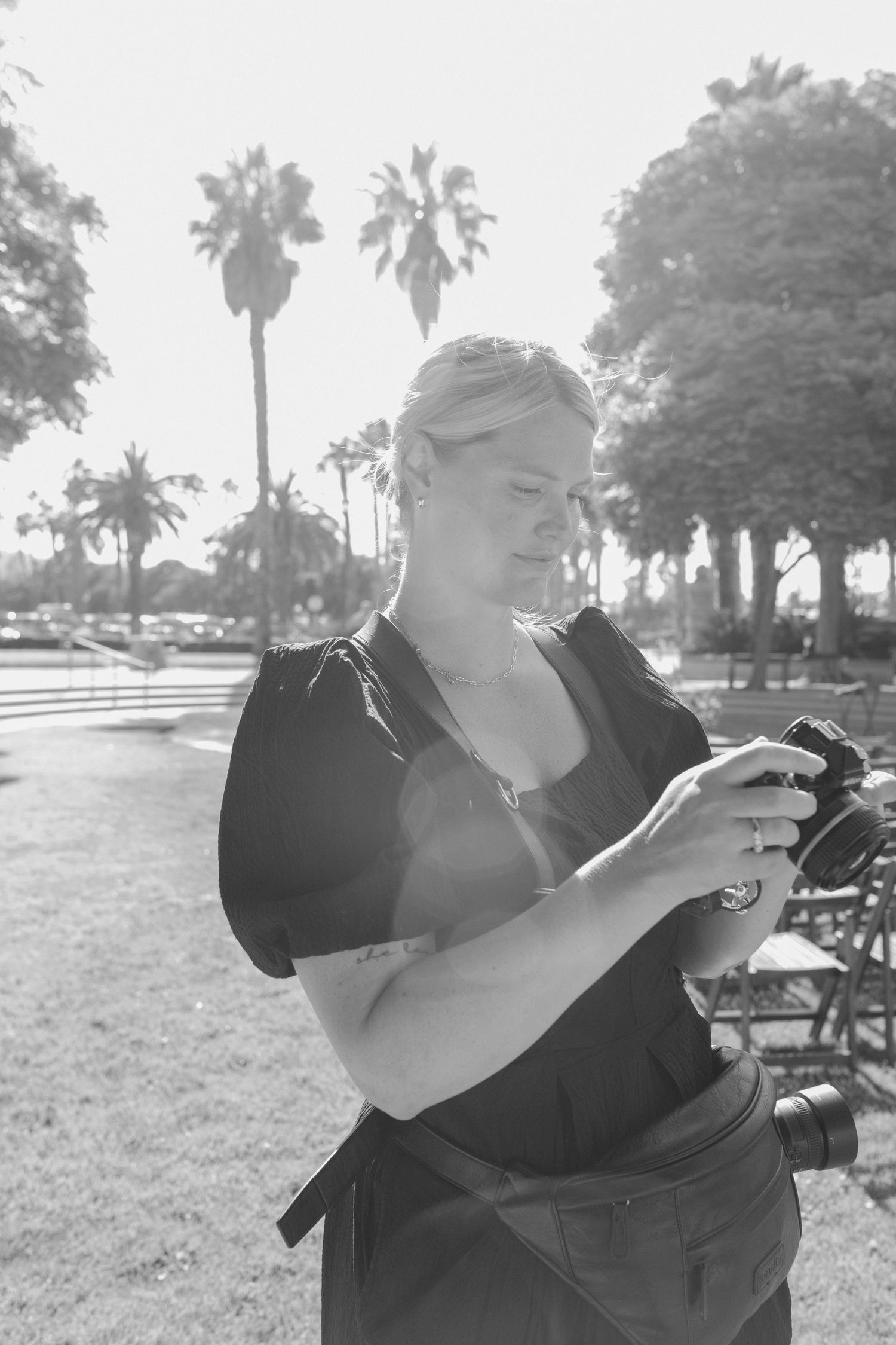 A person in black clothing takes photos outdoors with a professional camera in a sun-drenched setting.