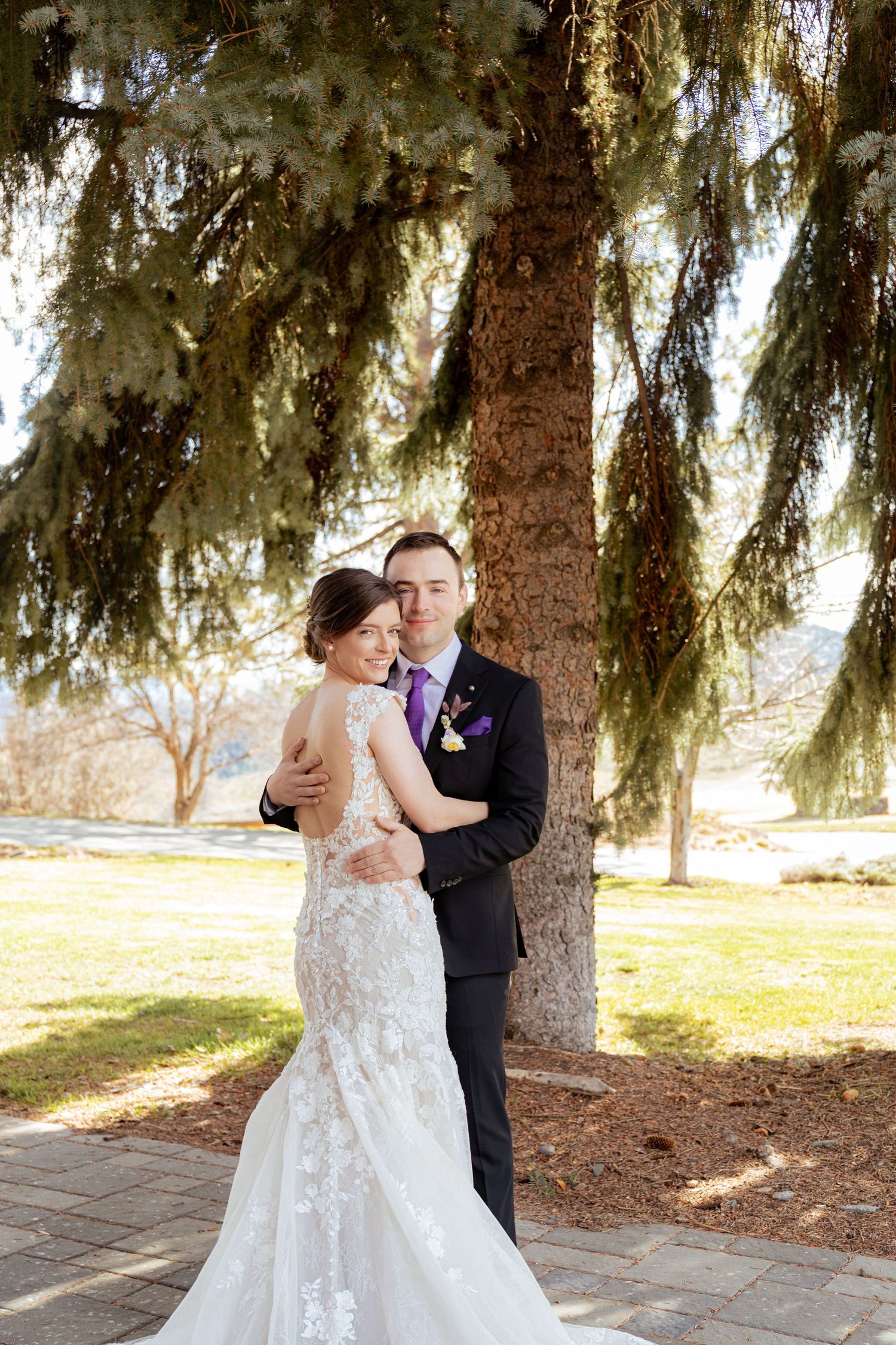 Romantic wedding couple embraces under evergreen trees in a park setting during their special day.