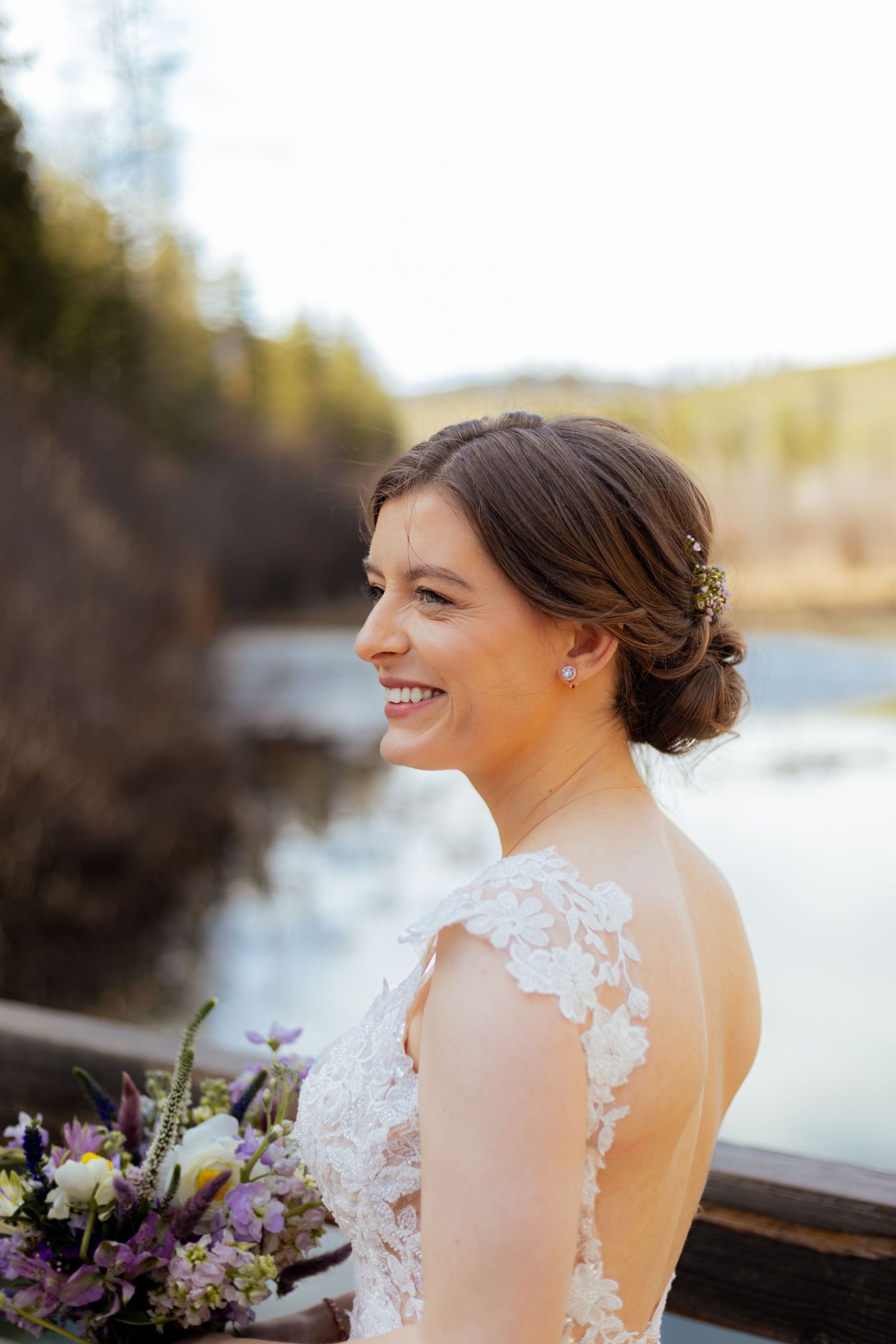 A bride in a lace wedding dress smiles joyfully by a waterfront during golden hour.