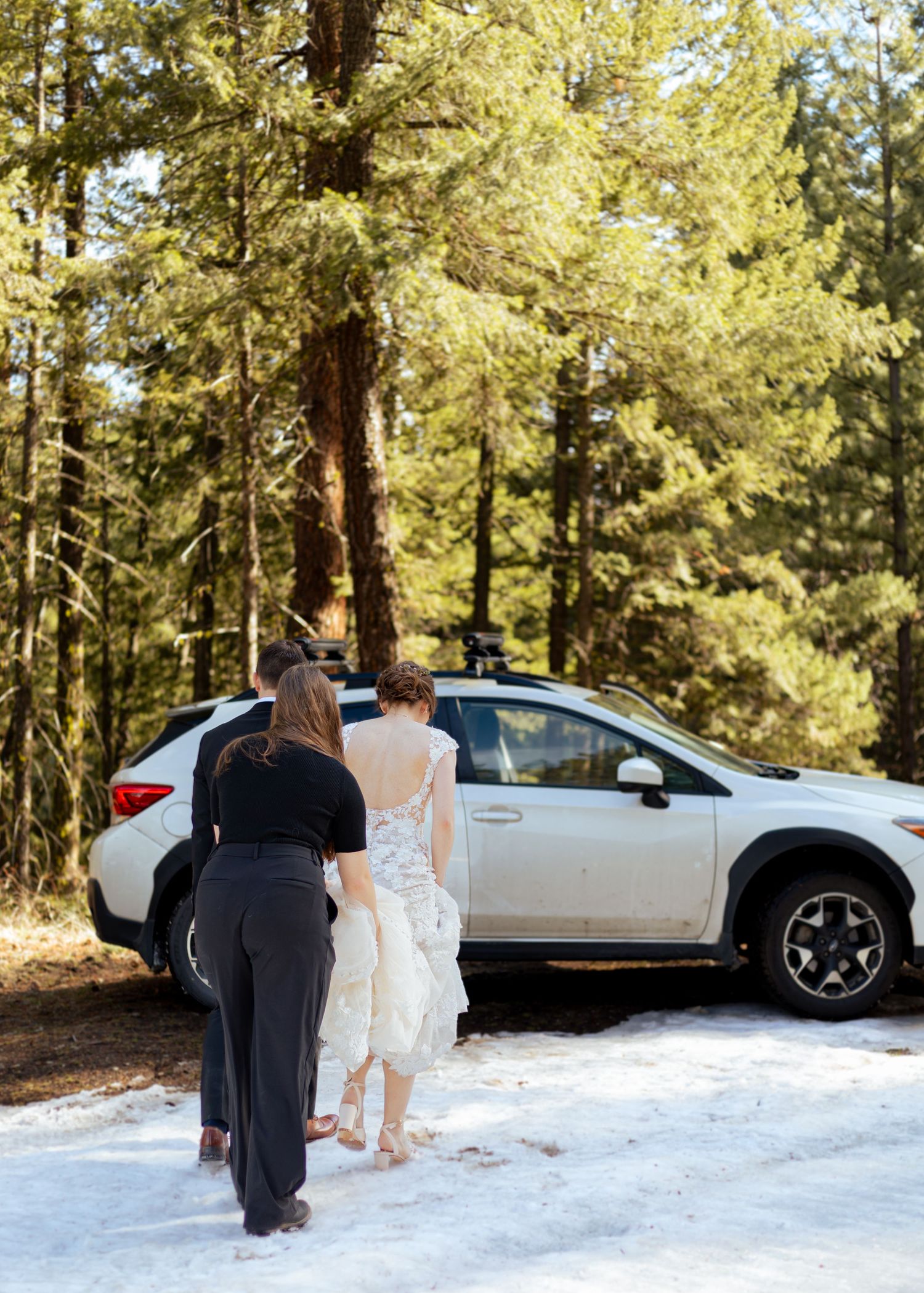 Two people walking in snowy forest with a white SUV parked among tall evergreen trees.