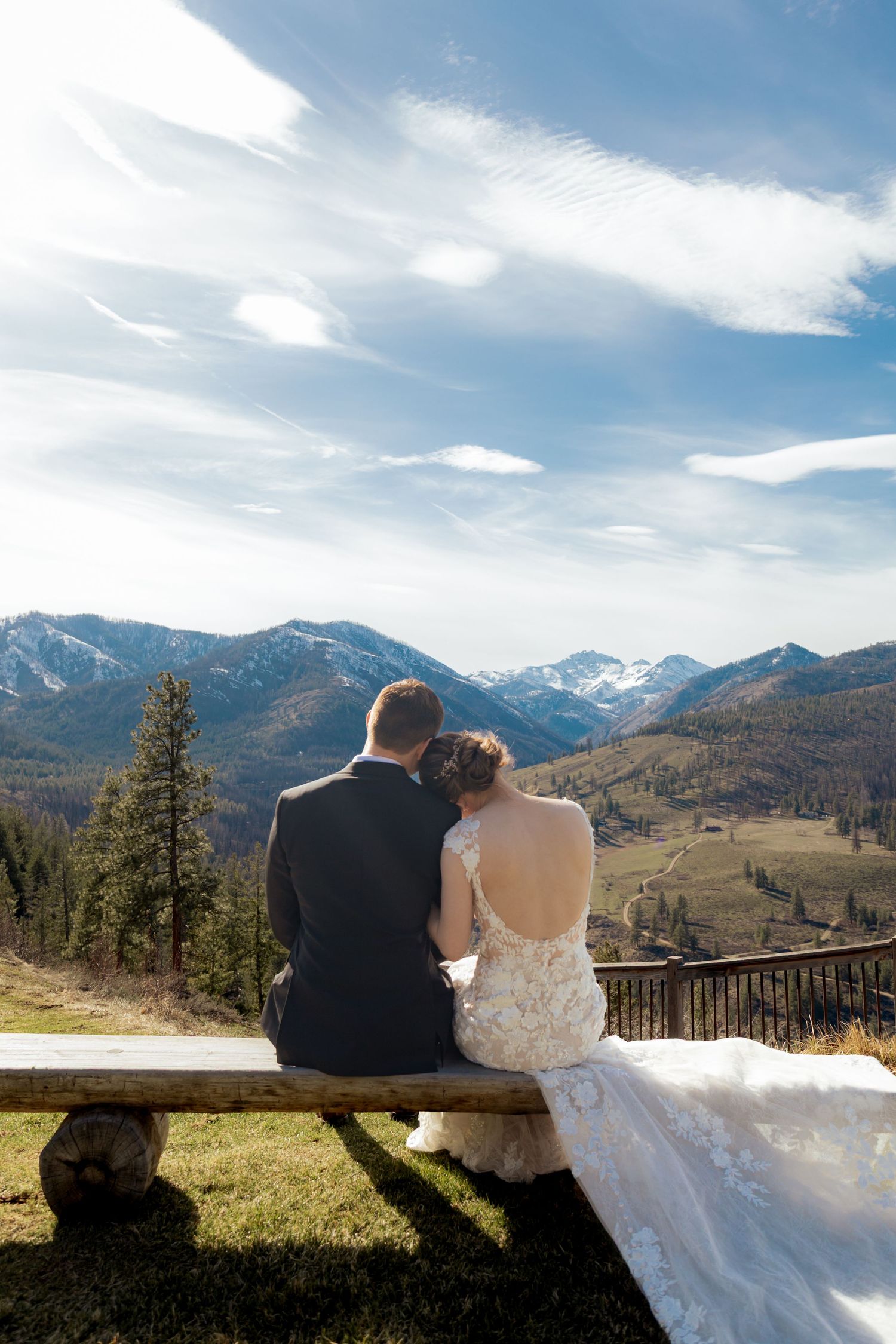 A couple sits on a rustic bench overlooking majestic snow-capped mountains in a scenic mountain wedding setting.