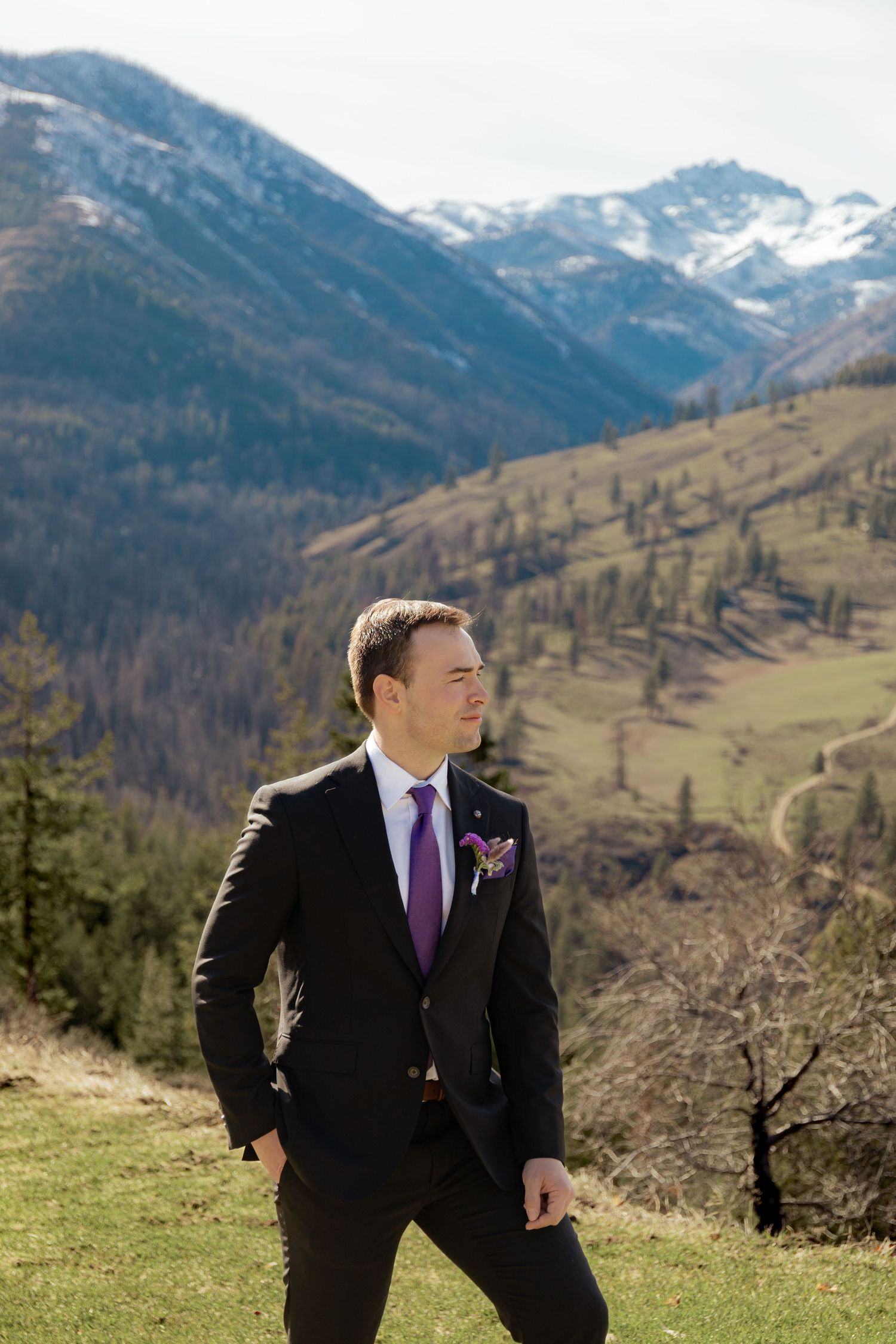 A man in a black suit and purple tie poses against a backdrop of dramatic mountain peaks and rolling hills.