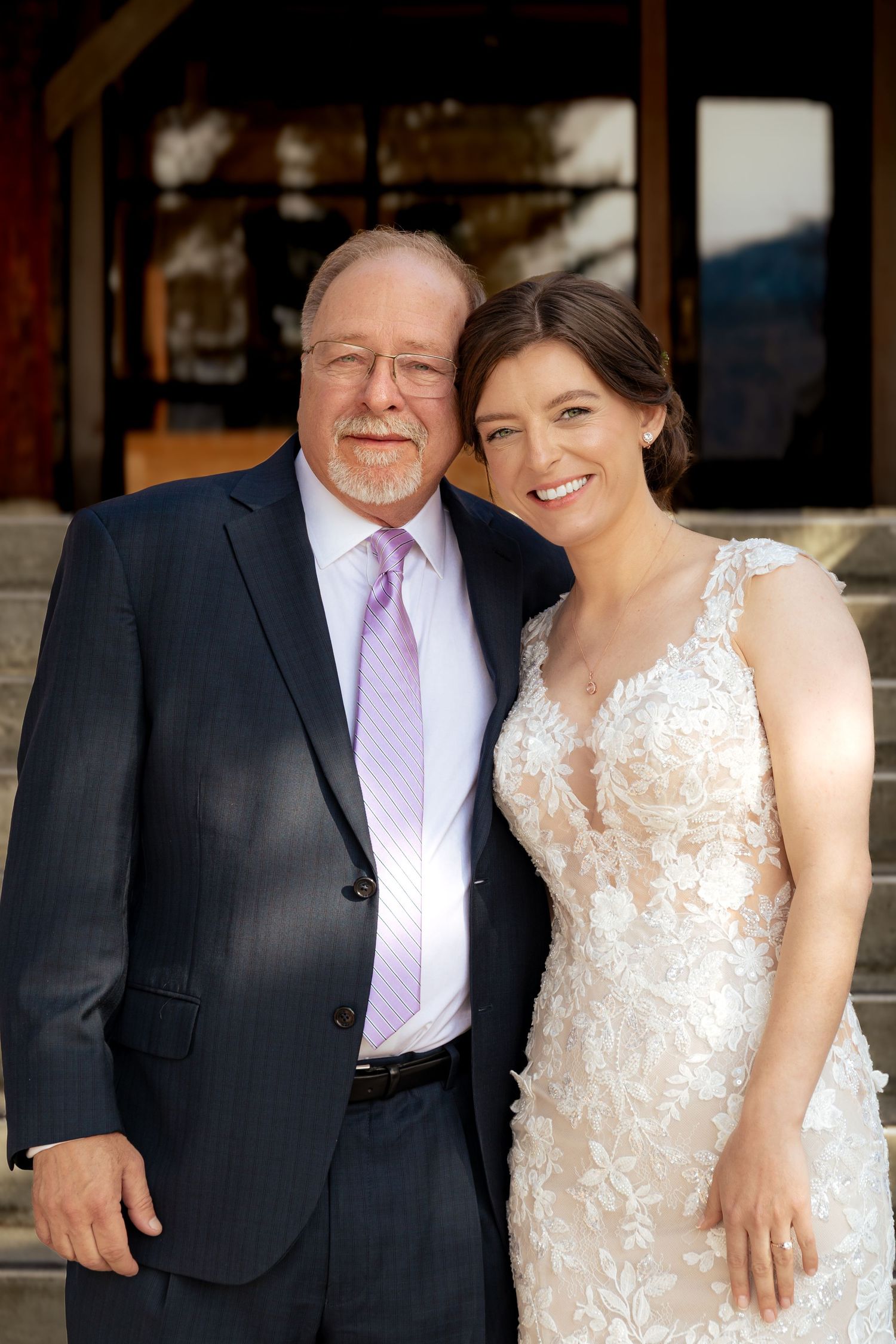 A formal portrait of a bride in a lace wedding dress standing with a guest in a dark suit and purple tie.