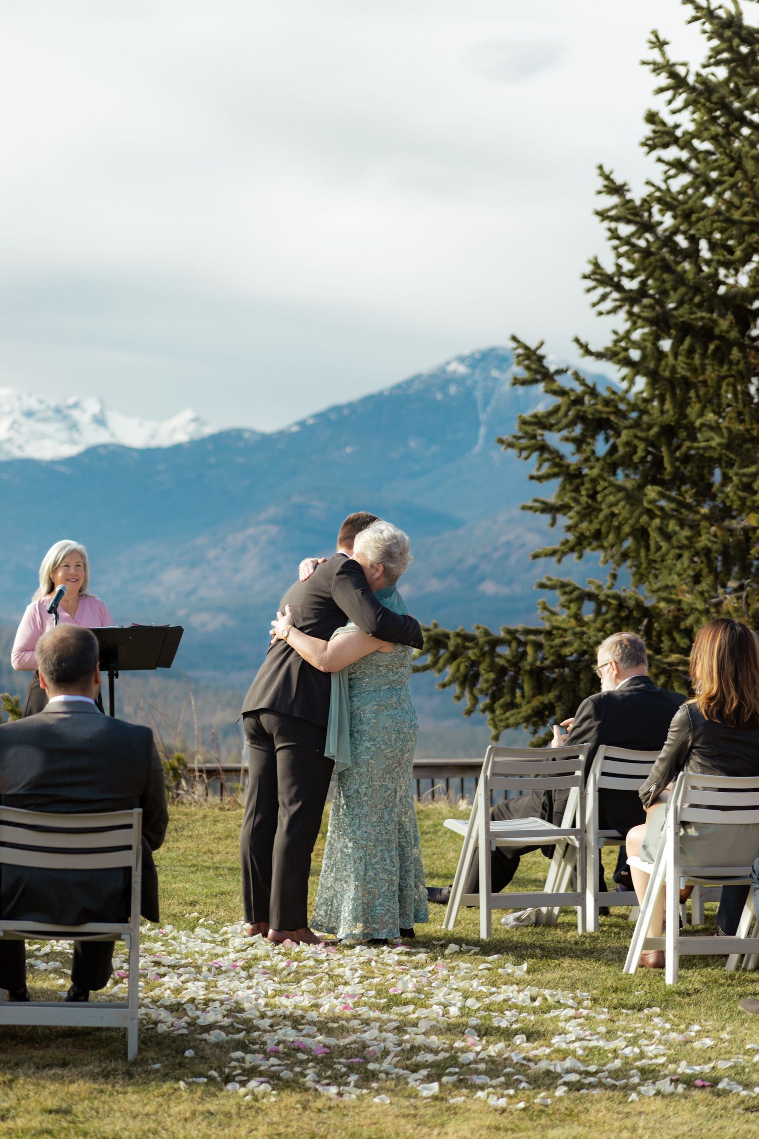People sitting in chairs overlooking a scenic mountain landscape with evergreen trees and snowy peaks in the distance.