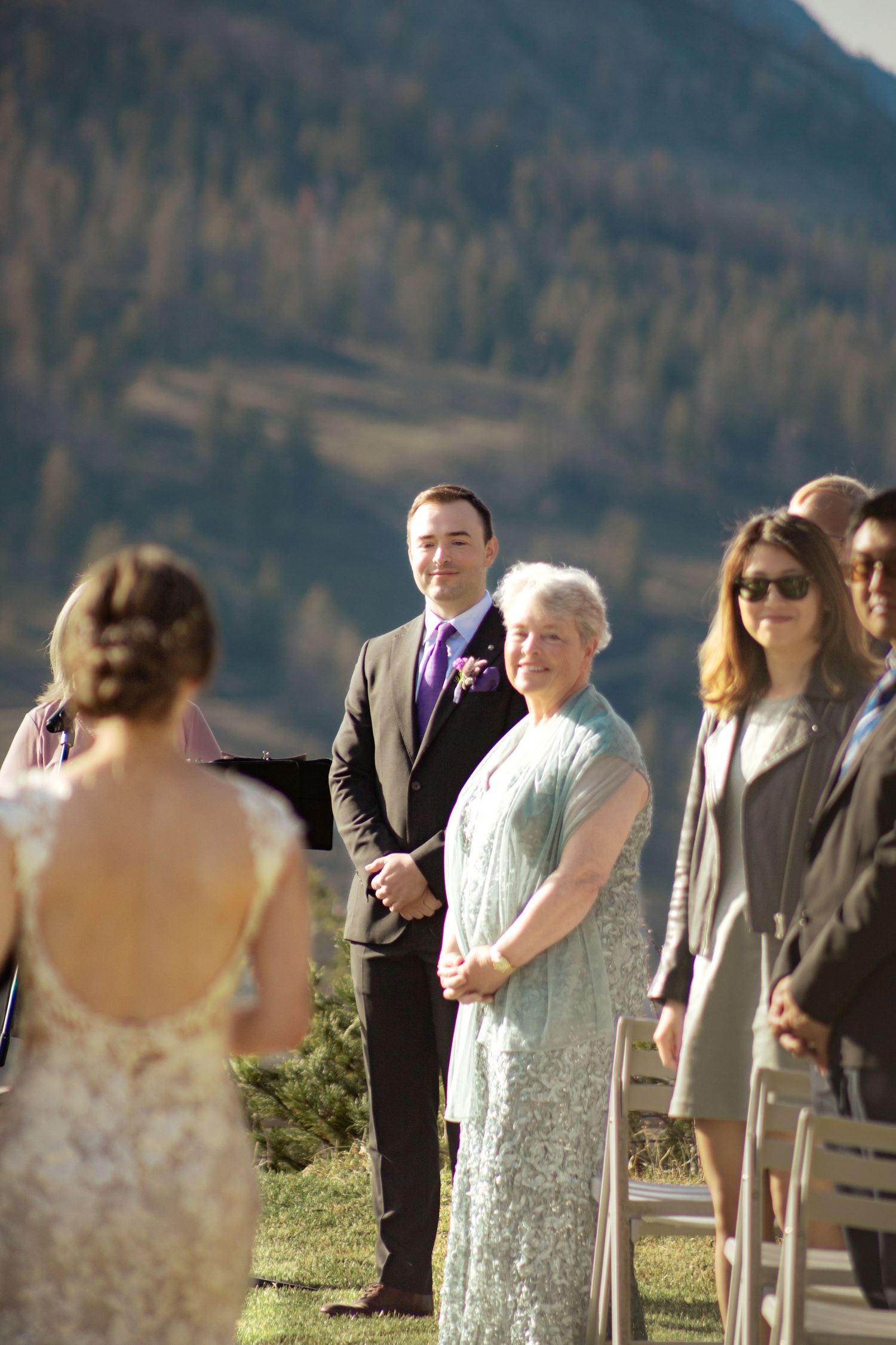 Wedding guests gather outdoors for a ceremony against a scenic mountain backdrop on a sunny day.