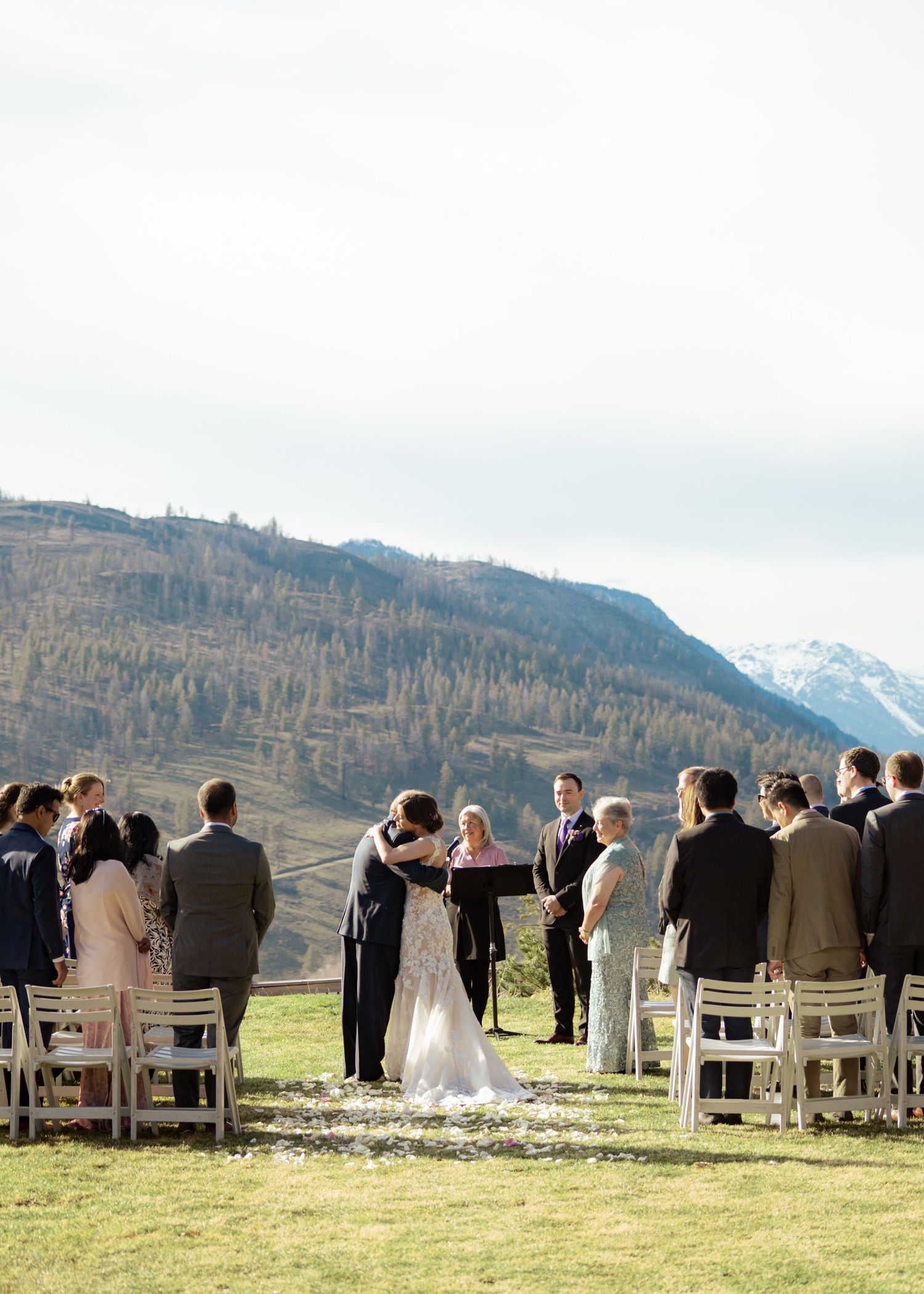 Wedding guests seated in white chairs attend an outdoor mountain ceremony with snow-capped peaks in the distance.
