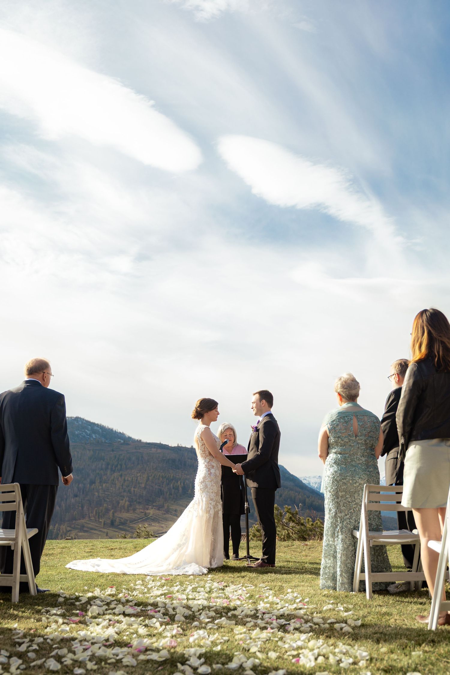 A scenic mountain wedding ceremony takes place outdoors with guests gathered on a grassy hilltop under blue skies.