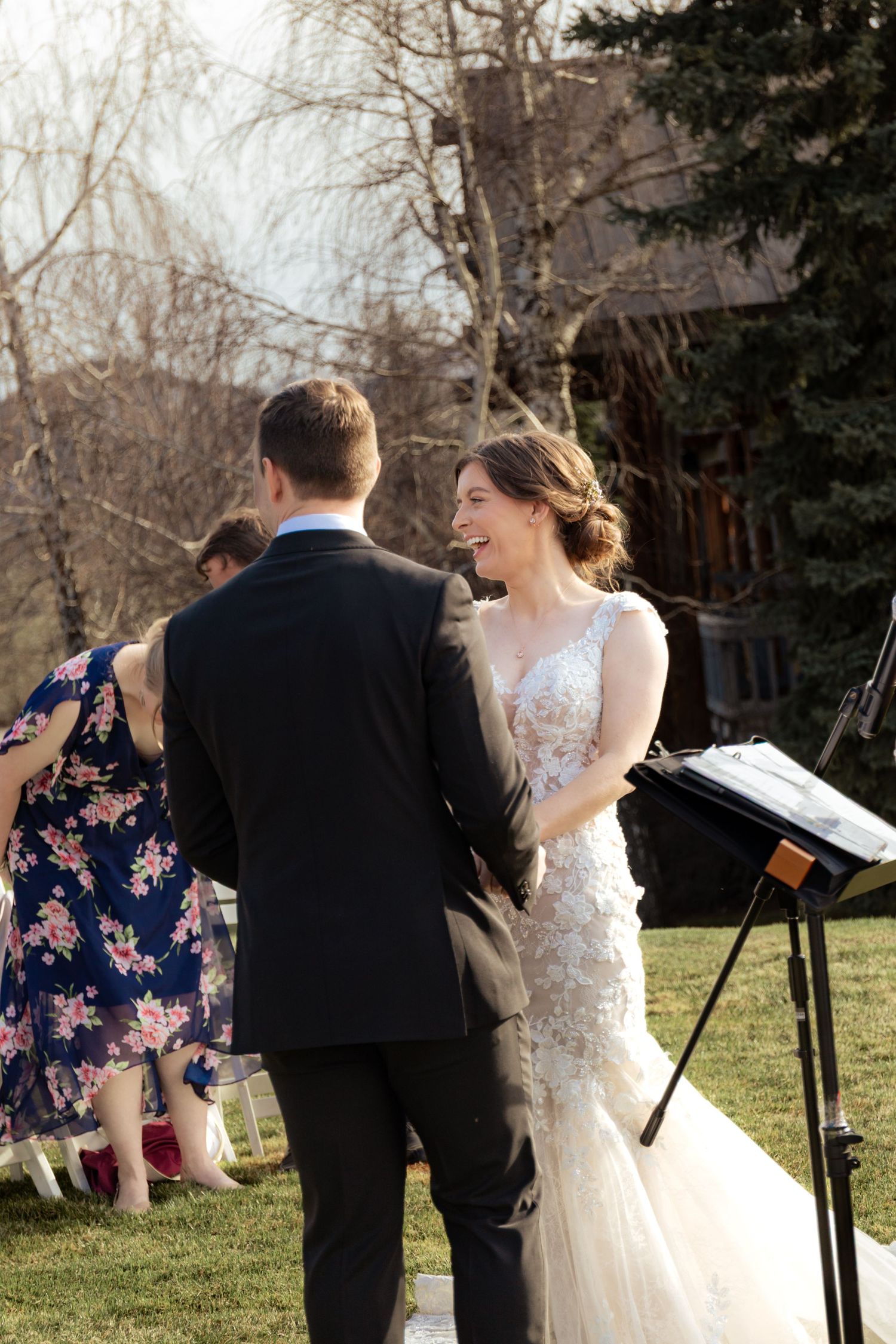 An outdoor wedding ceremony takes place in a garden setting with guests gathered on a sunny winter afternoon.