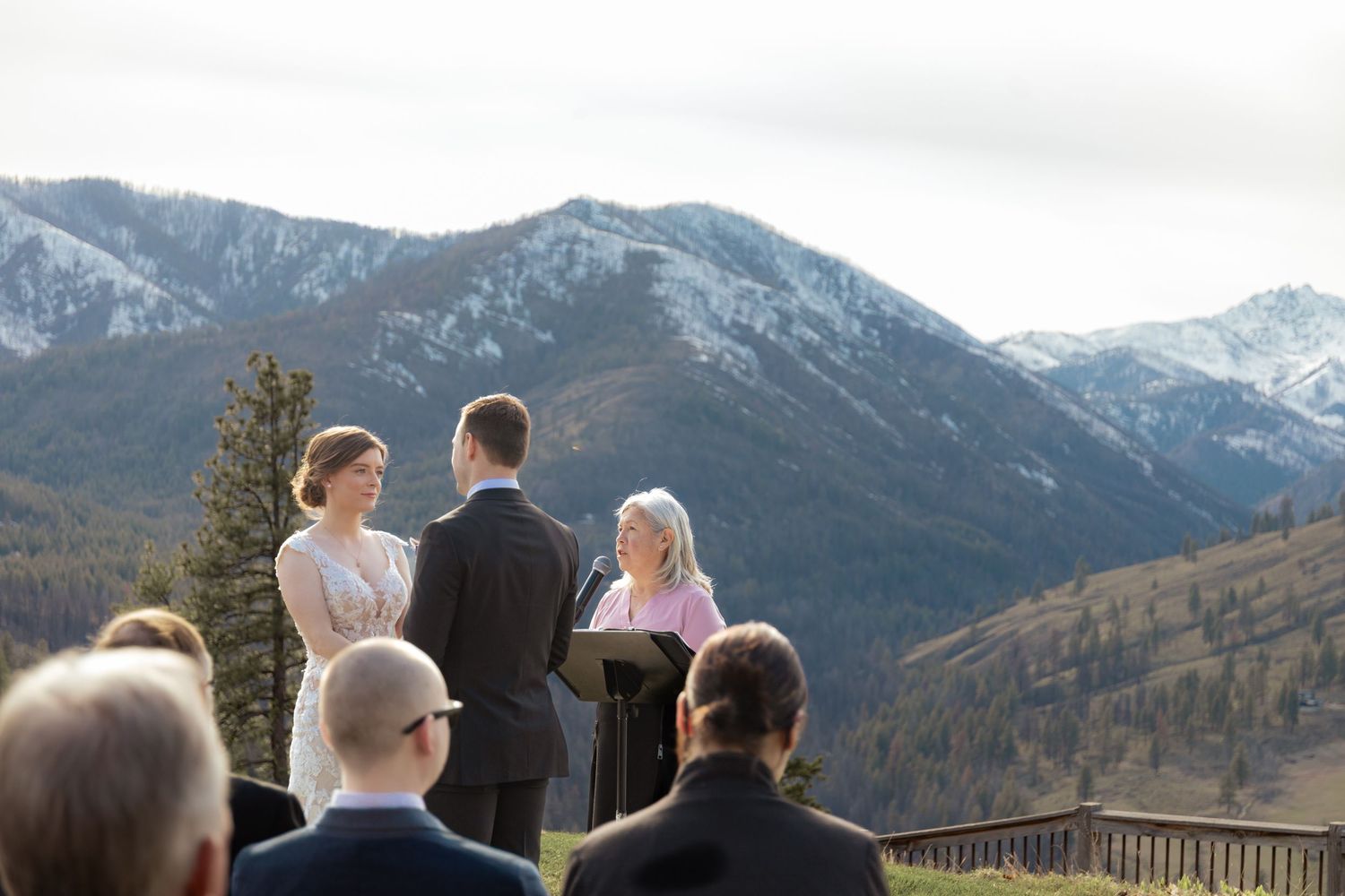 A wedding ceremony takes place on a scenic mountain overlook with snow-capped peaks in the background.