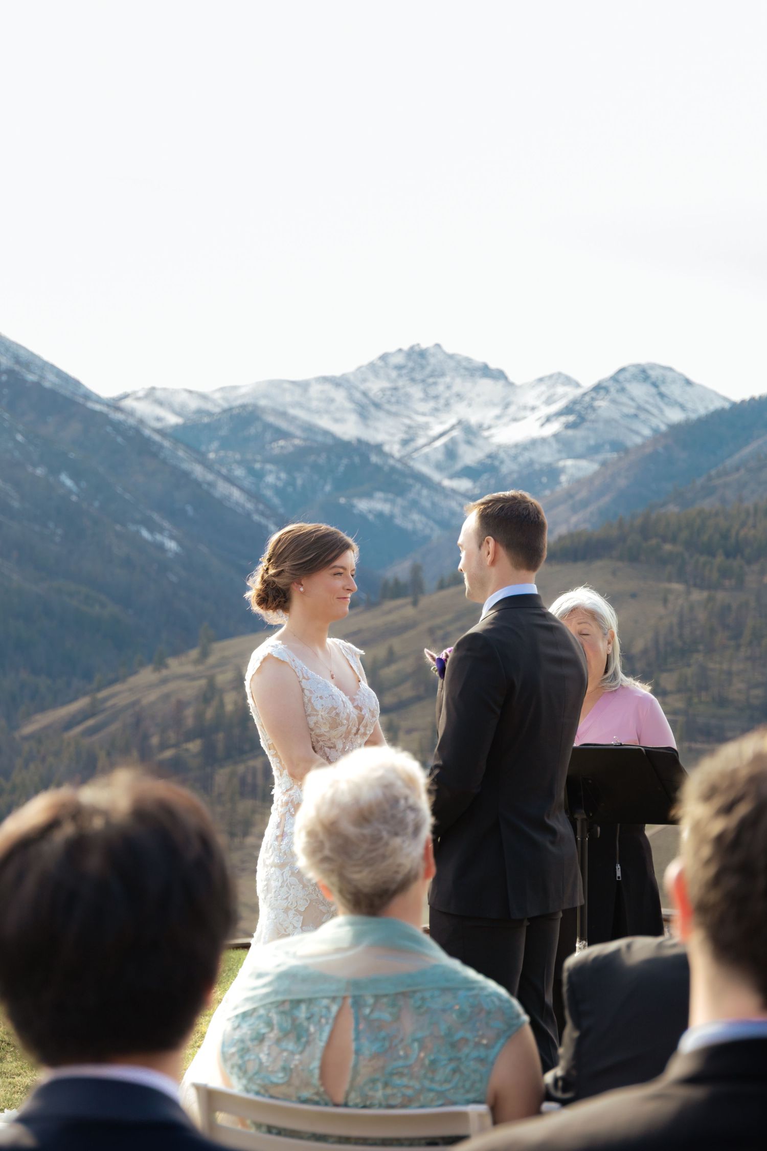 A wedding ceremony takes place in front of snow-capped mountain peaks with guests watching the couple exchange vows.