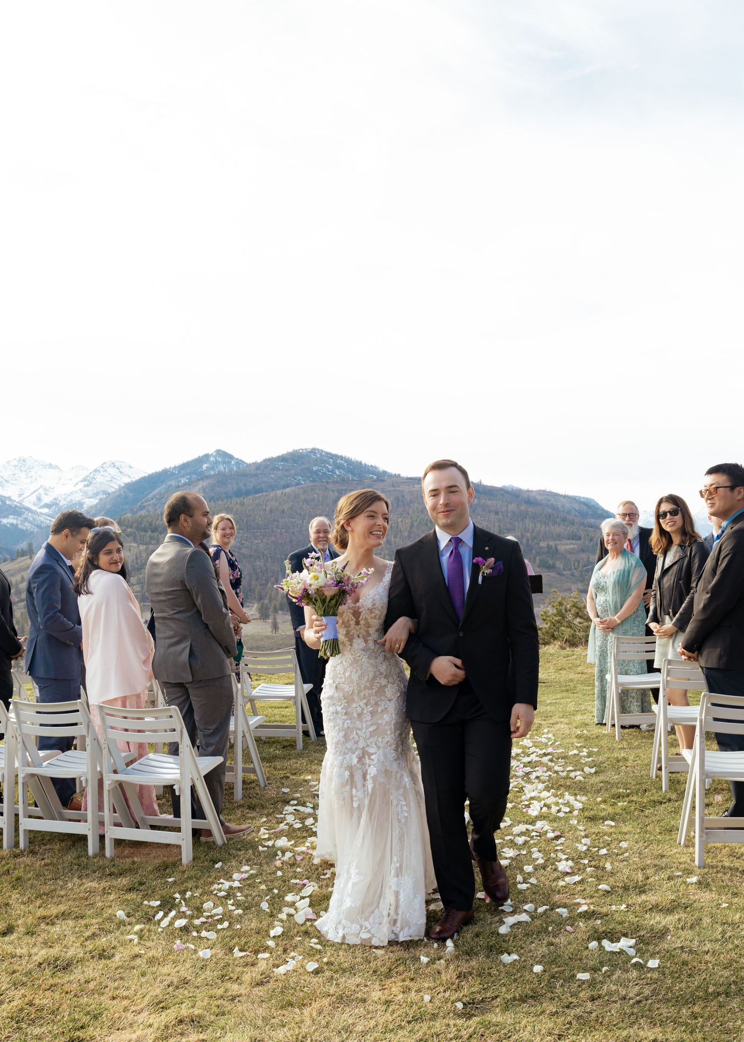A panoramic view of a wedding ceremony on a grassy hillside with mountain peaks in the background and scattered flower petals.