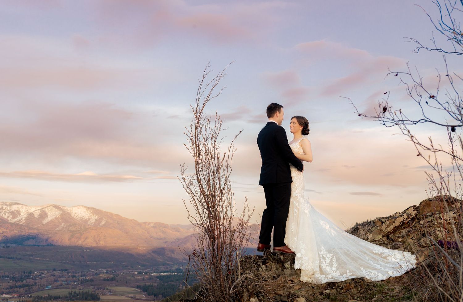 A couple in wedding attire stands on a mountain overlook at sunset with snow-capped peaks in the distance.