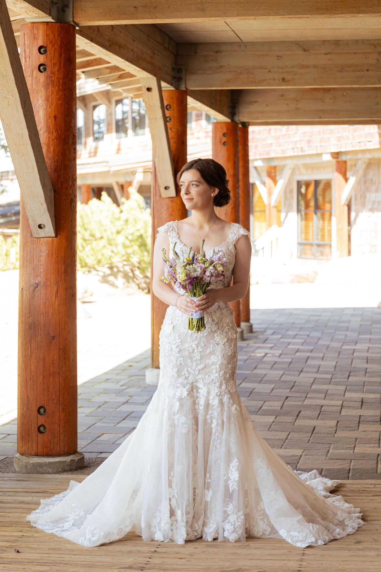 A bride in a lace wedding gown poses under wooden beams in an outdoor covered walkway with natural lighting.