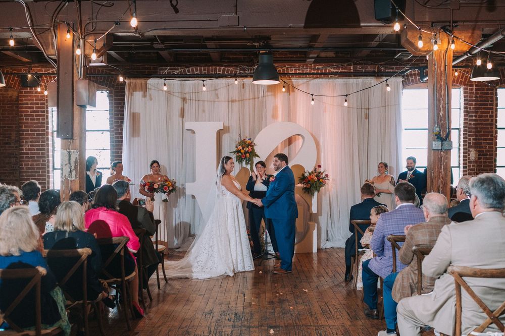 Wedding guests mingle and dance in a rustic brick-walled reception venue with exposed beams and string lights.