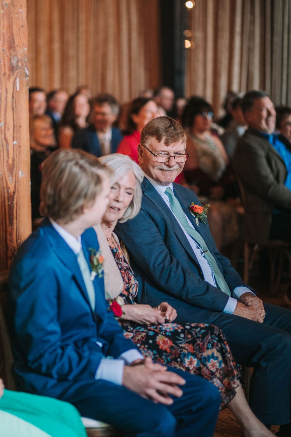 Wedding guests seated in wooden chairs smile and laugh during an indoor ceremony.