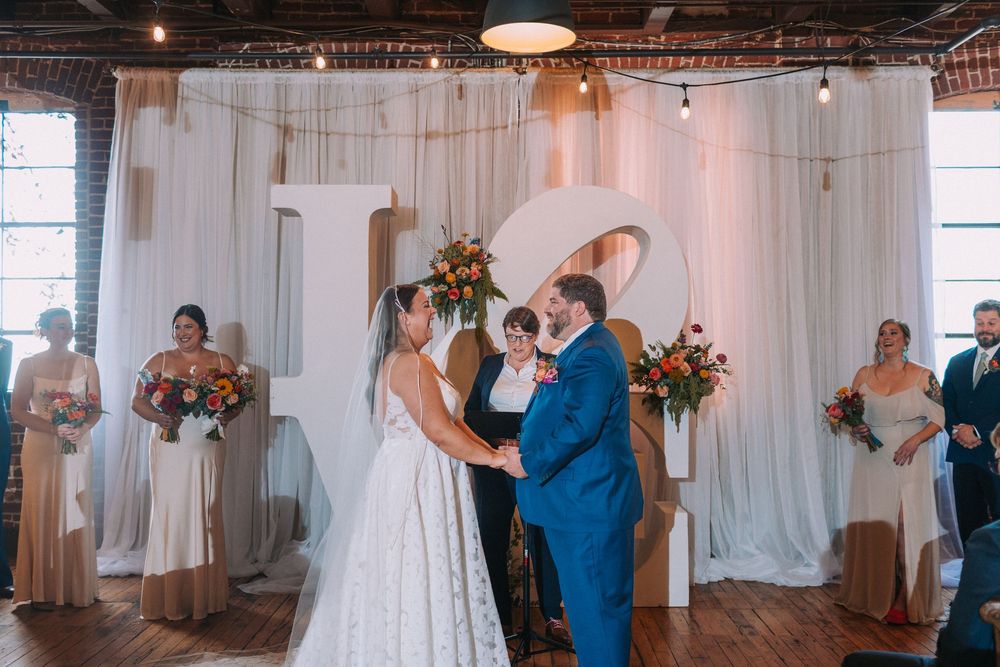 A wedding ceremony takes place under white drapes with floral arrangements in an industrial-style venue.