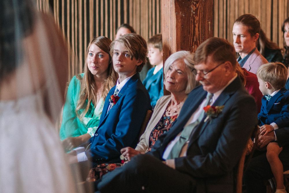 Wedding guests seated in rows watch attentively during an indoor ceremony.