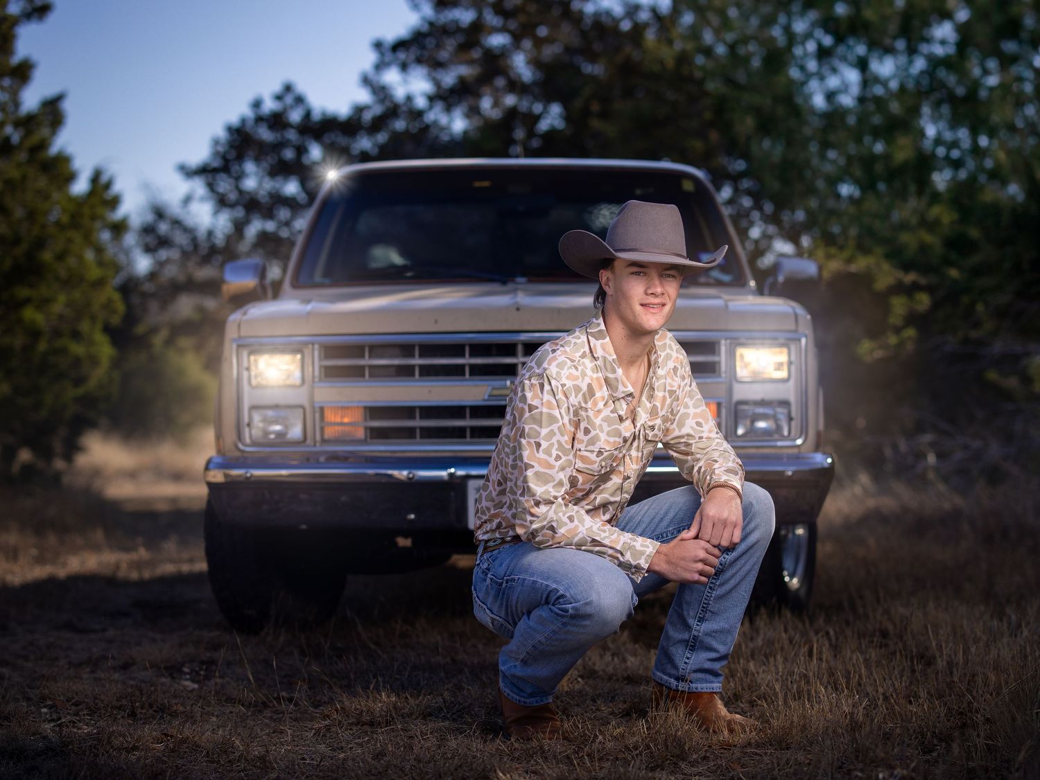 A young man in a camouflage  shirt and jeans poses by a vintage pickup truck in a rustic outdoor setting at sunset.