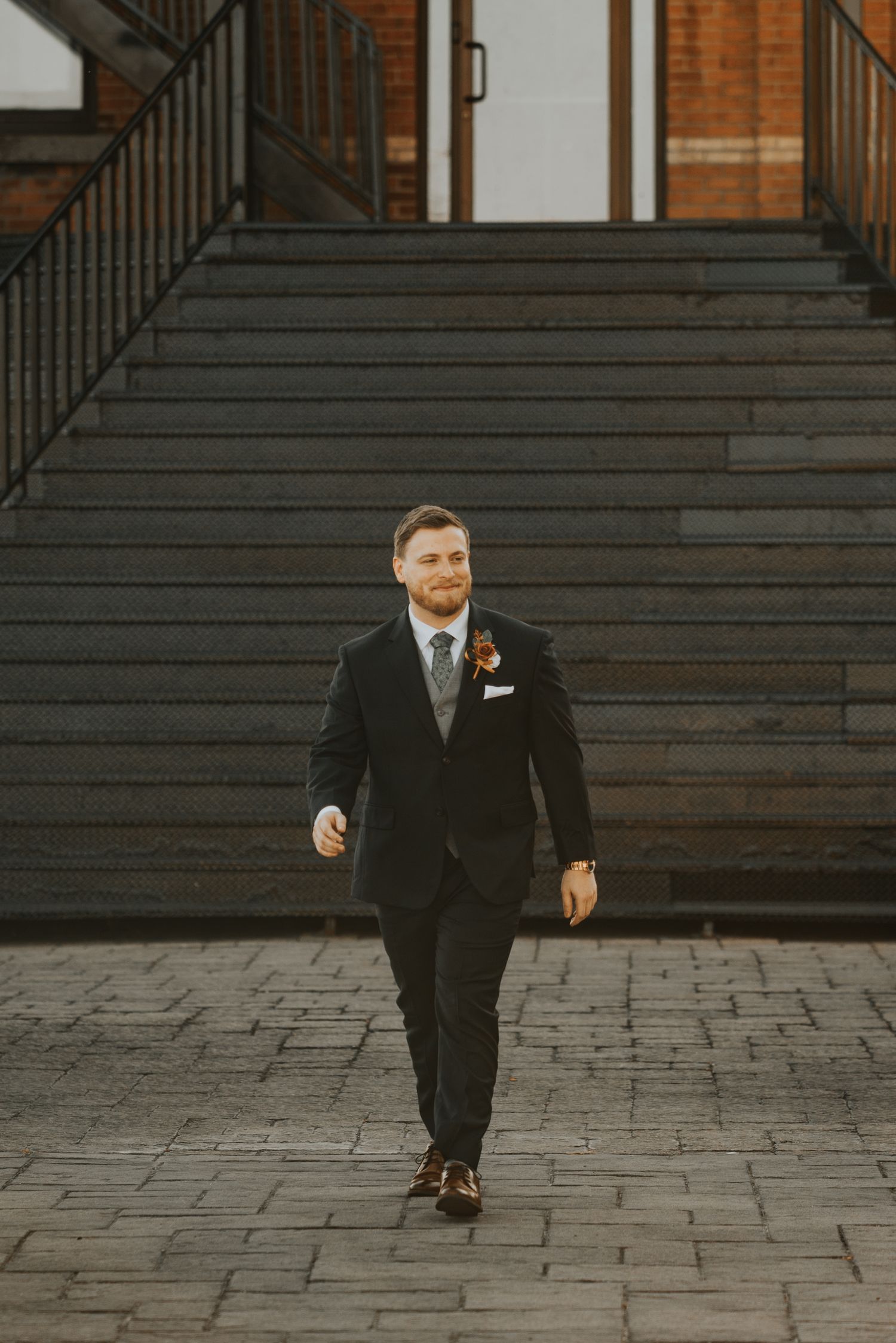 A groom in a black tuxedo walks down stone steps in front of a brick building during a wedding photo session.