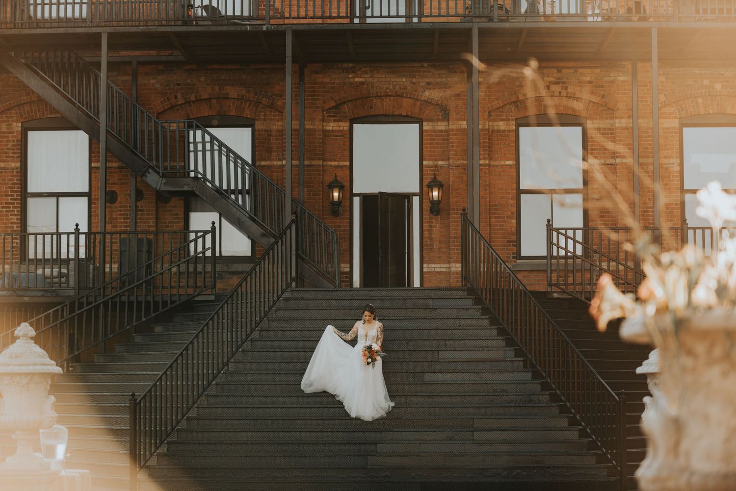 Couple in wedding attire standing on steps of historic brick building with fire escapes at sunset.
