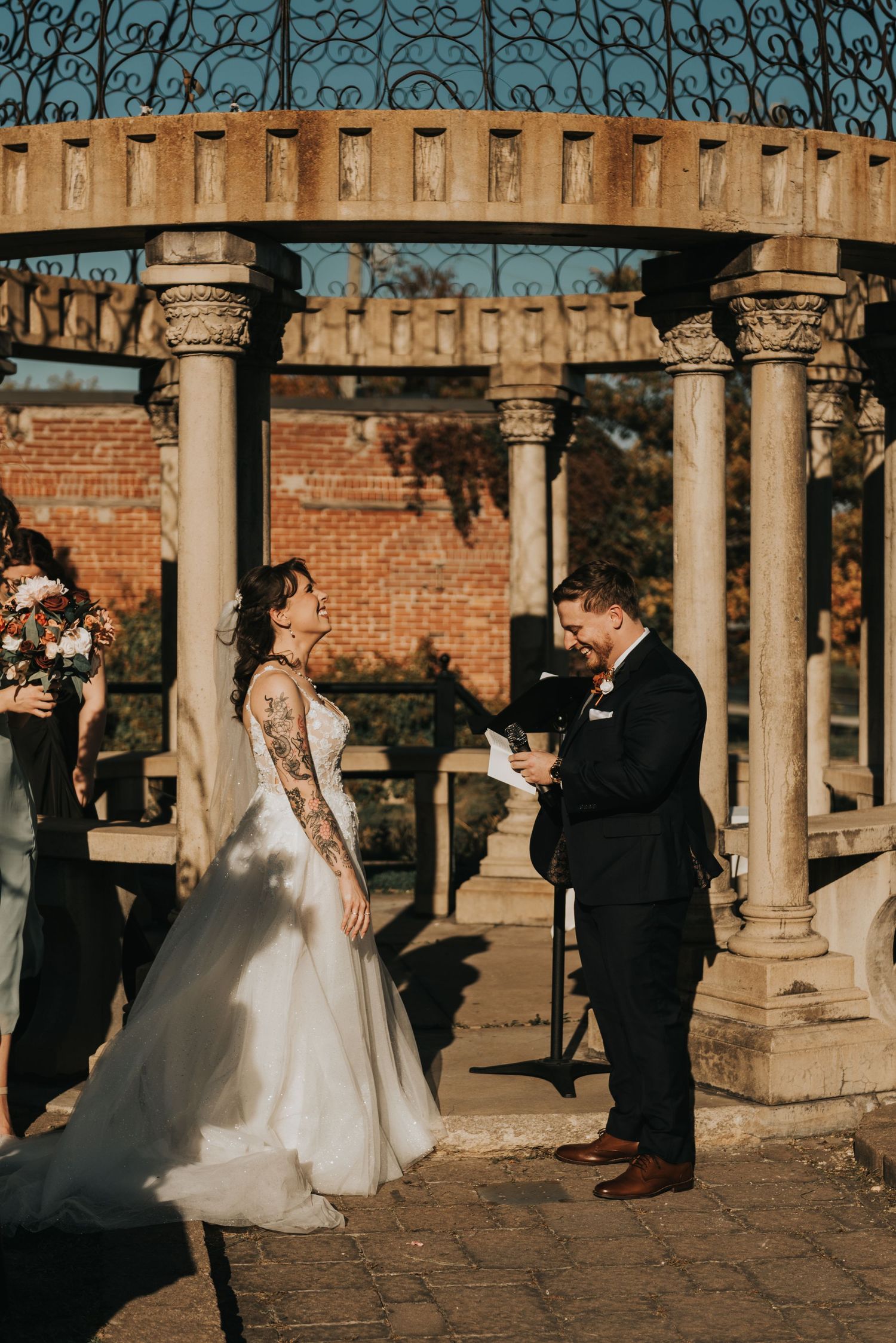 A wedding ceremony takes place under a columned pavilion with brick walls on a sunny afternoon.