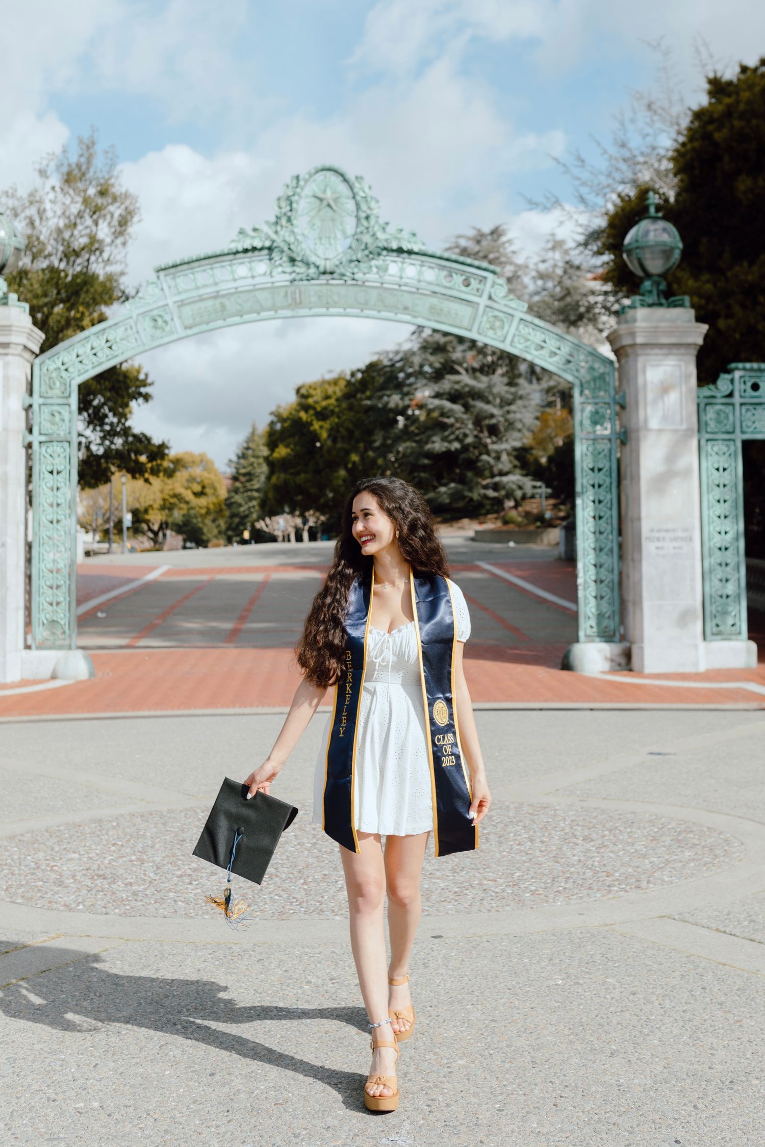 Student walks through iconic Sather Gate entrance at UC Berkeley campus on a sunny day for her grad photoshoot. She wears her sash and cap.