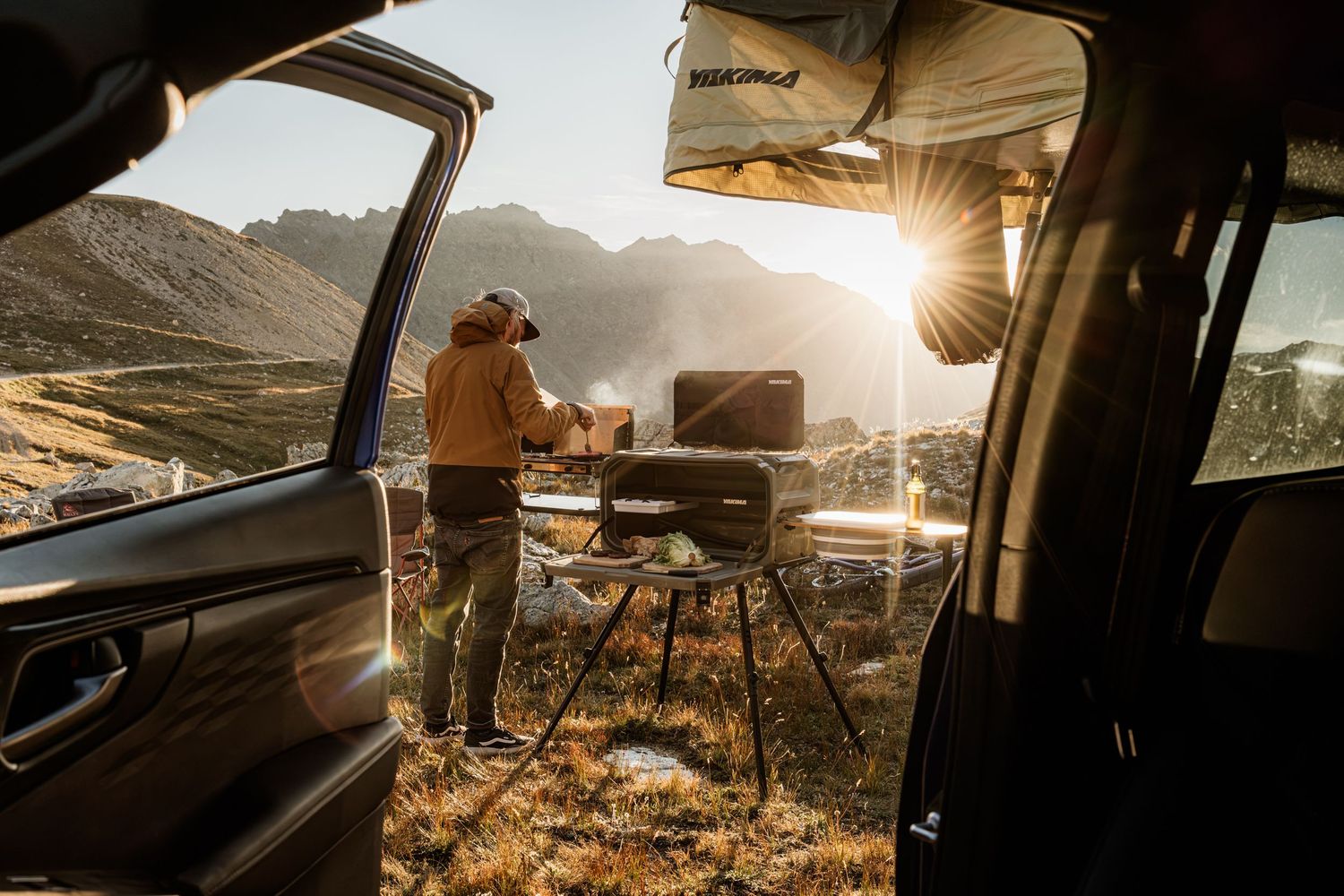 Person camping in car with scenic mountain view at sunset while enjoying coffee from thermos.