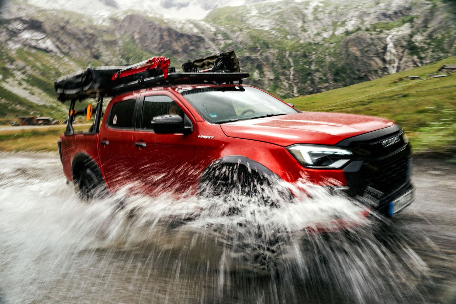 Red Isuzu D-Max splashes through water while driving on a mountain road in dramatic action sequence.