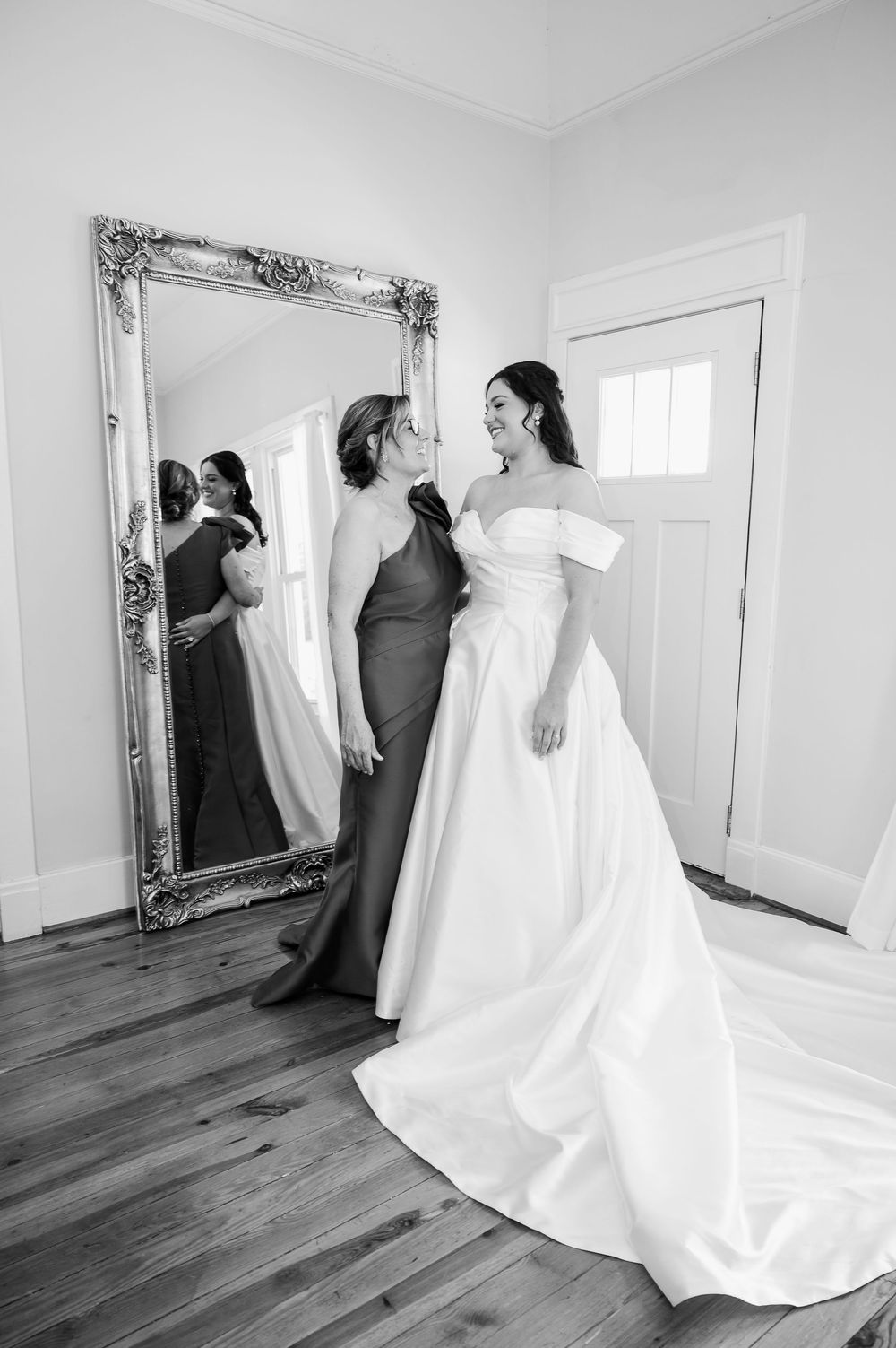 Black and white series showing a wedding dress being admired in an elegant room with hardwood floors and ornate mirror.