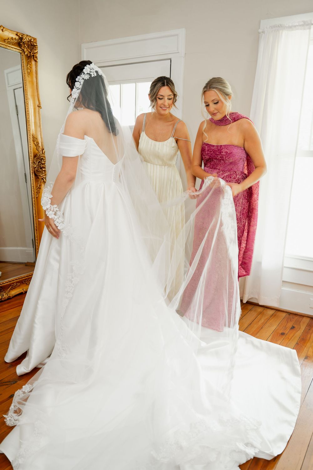 Wedding dress preparation scene with flowing train and veil being arranged on hardwood floor.