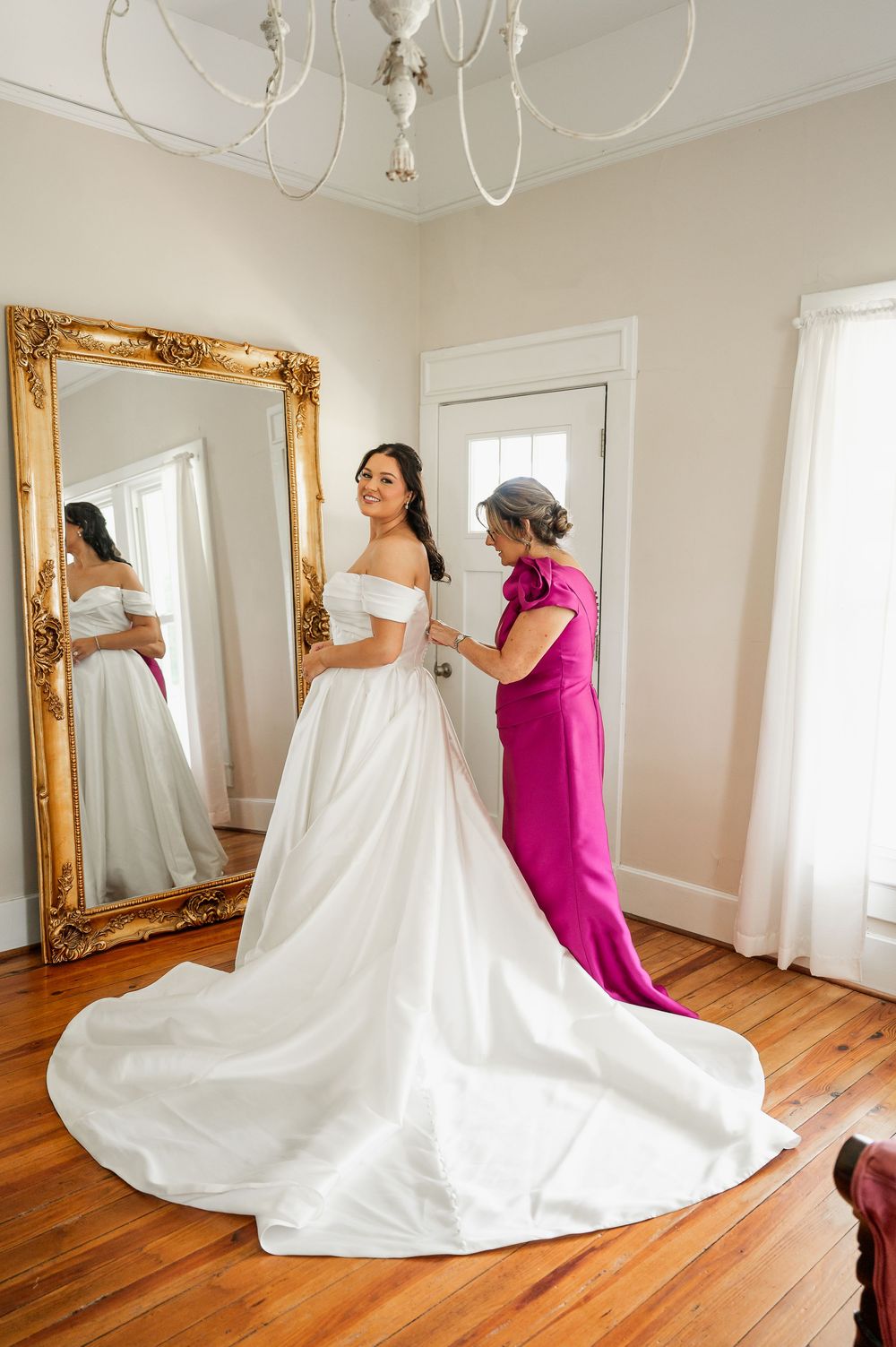 Wedding dress preparation scene with elegant white gown and attendant in bright pink in natural light.