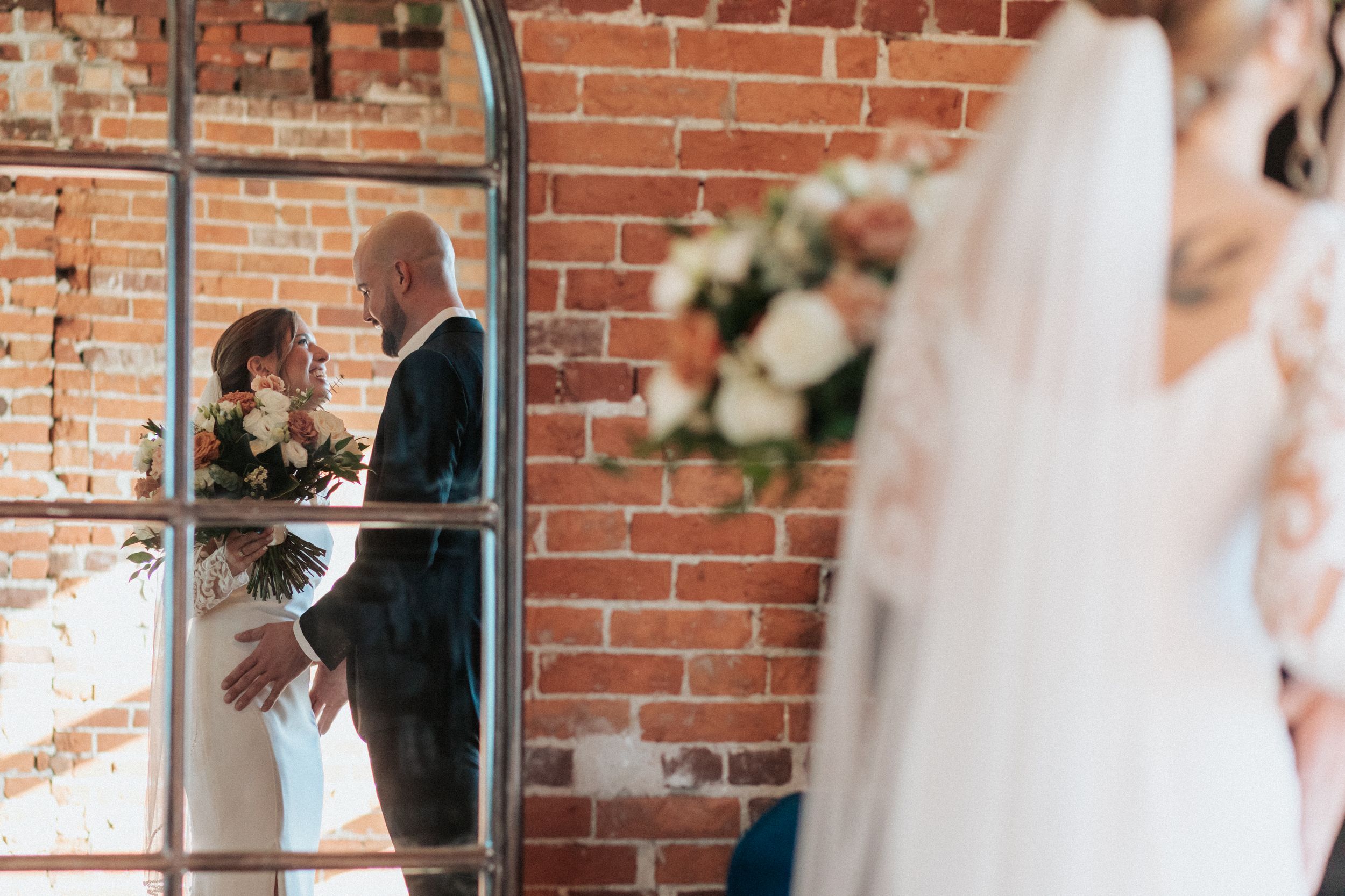 First look wedding moment captured against rustic brick backdrop with window light.
