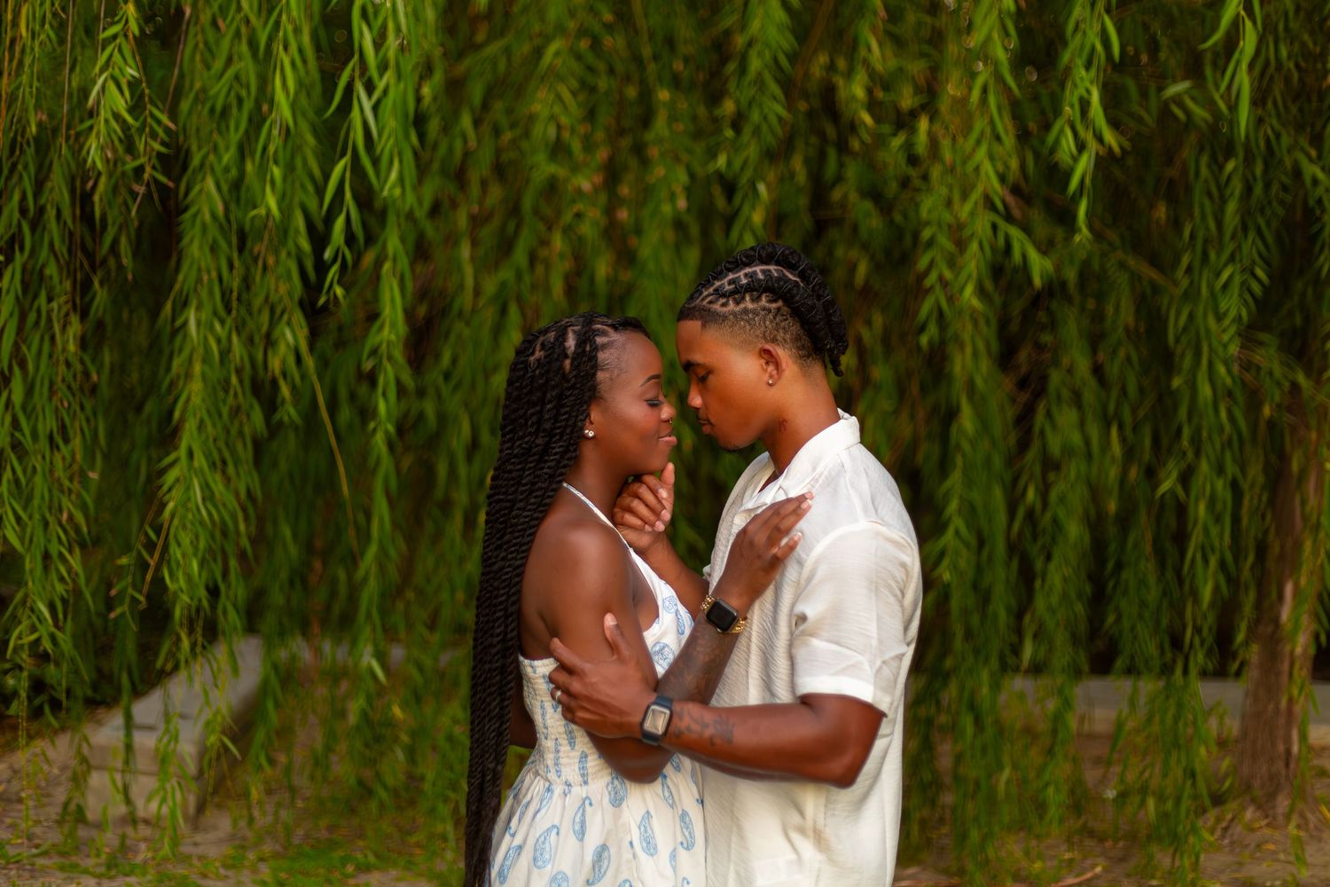 Romantic couple embraces under cascading willow branches during outdoor photo session in natural setting.