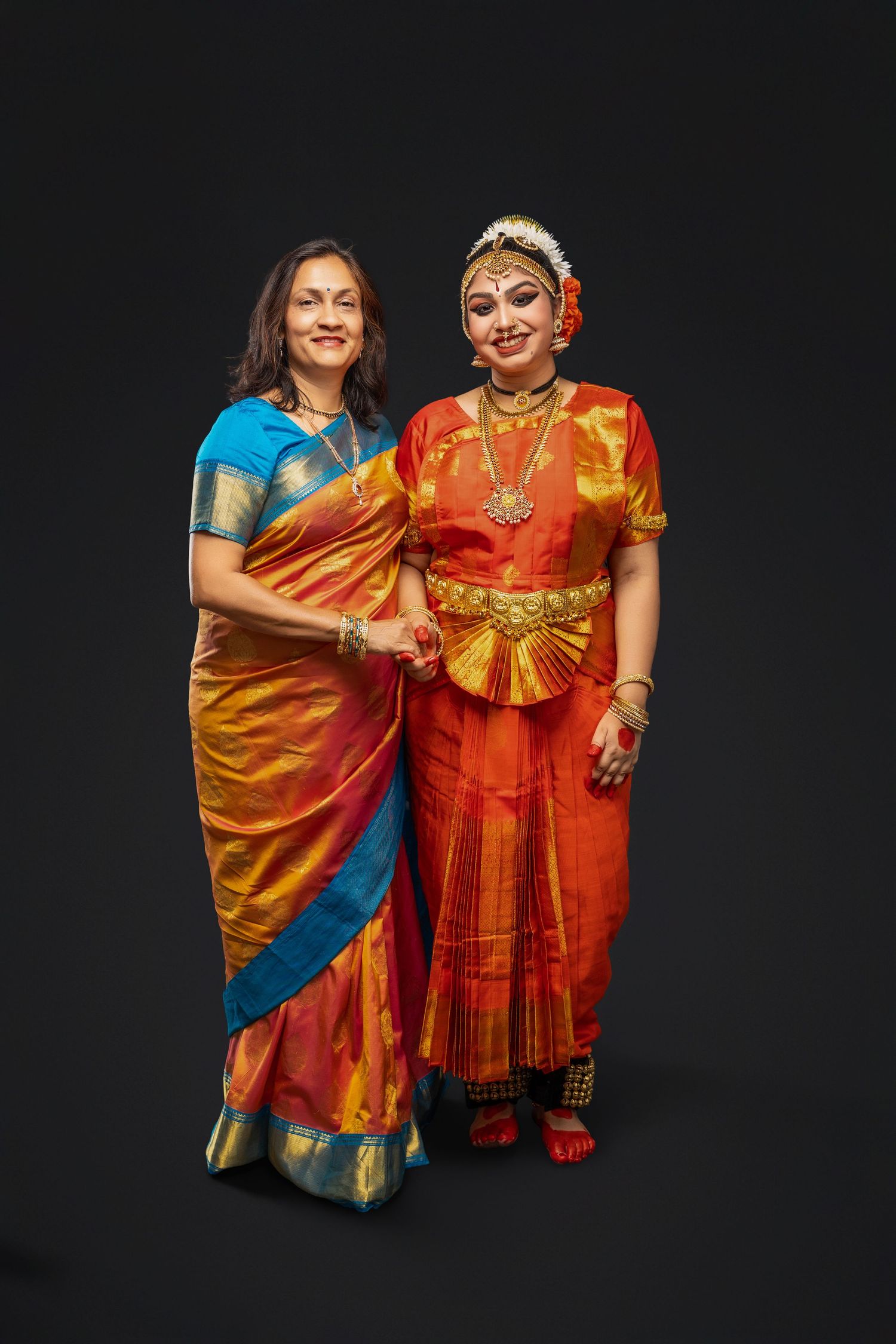 Two dancers perform traditional Indian classical dance moves in vibrant orange and gold costumes against a black background.