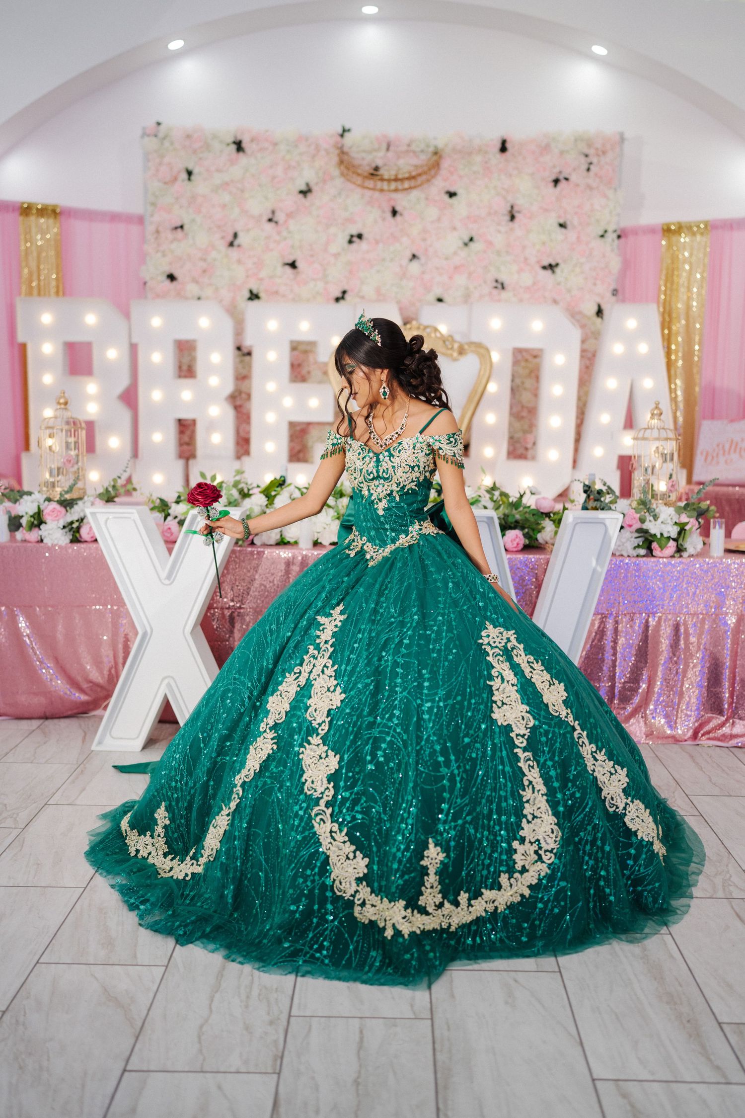 Elegant emerald green quinceañera ball gown with gold lace appliques sparkles in a pink decorated celebration venue.