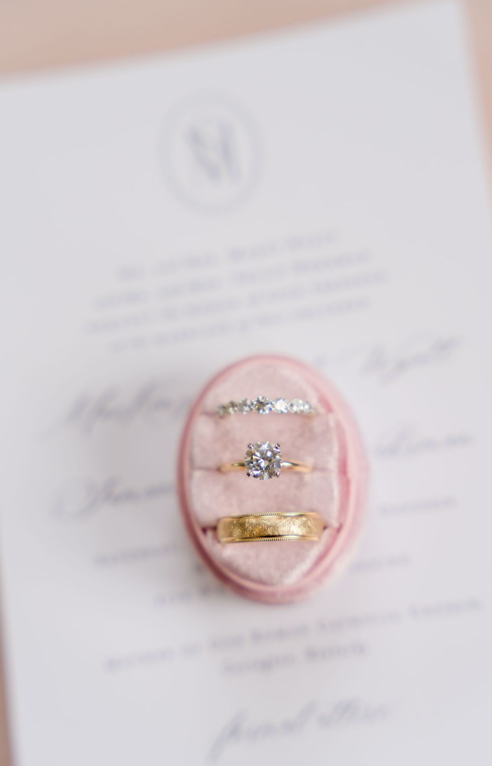 Wedding rings displayed on pink velvet ring box with invitation details in background.